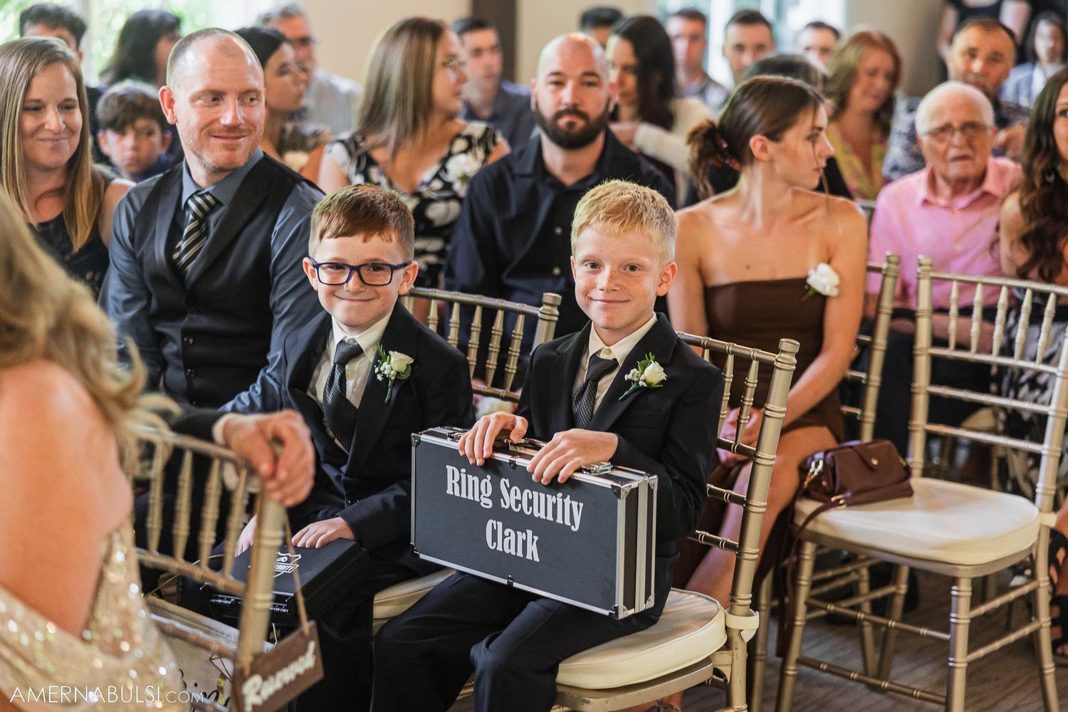 Young ring bearers hold a decorative sign while seated in gold chairs during wedding ceremony.