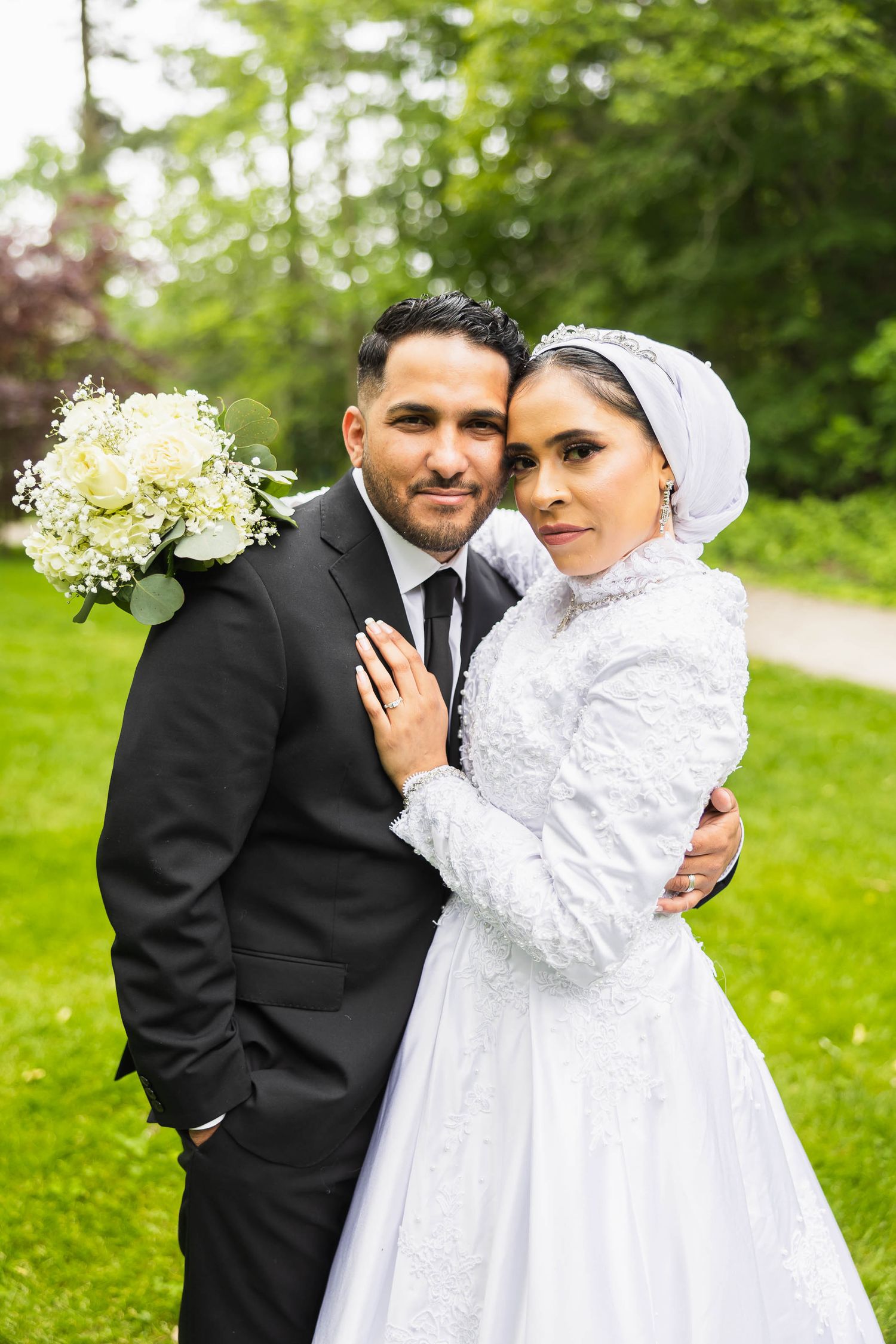 A bride in white lace and hijab poses with her groom on their wedding day.