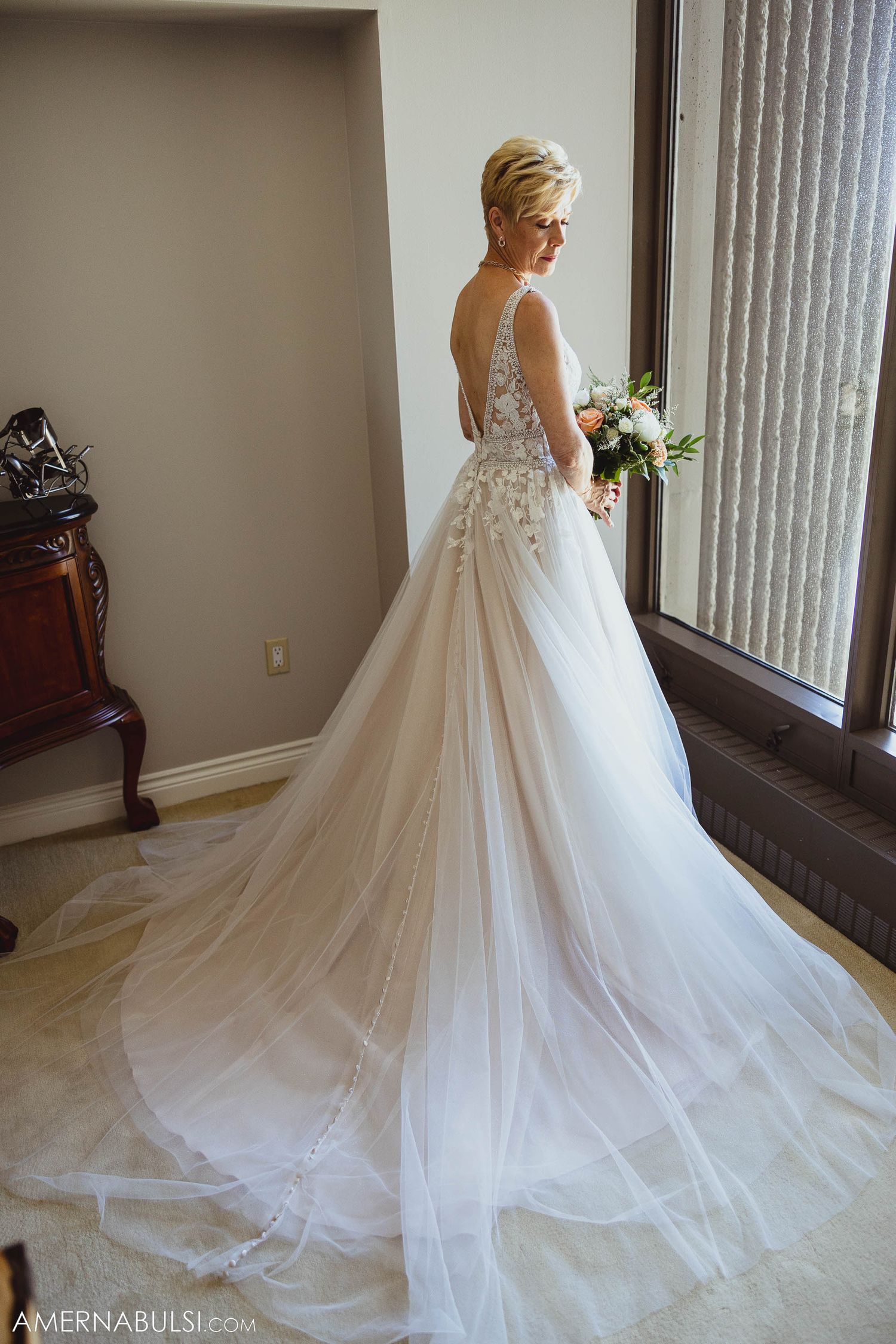 Elegant bride poses in flowing wedding gown with dramatic train near window.