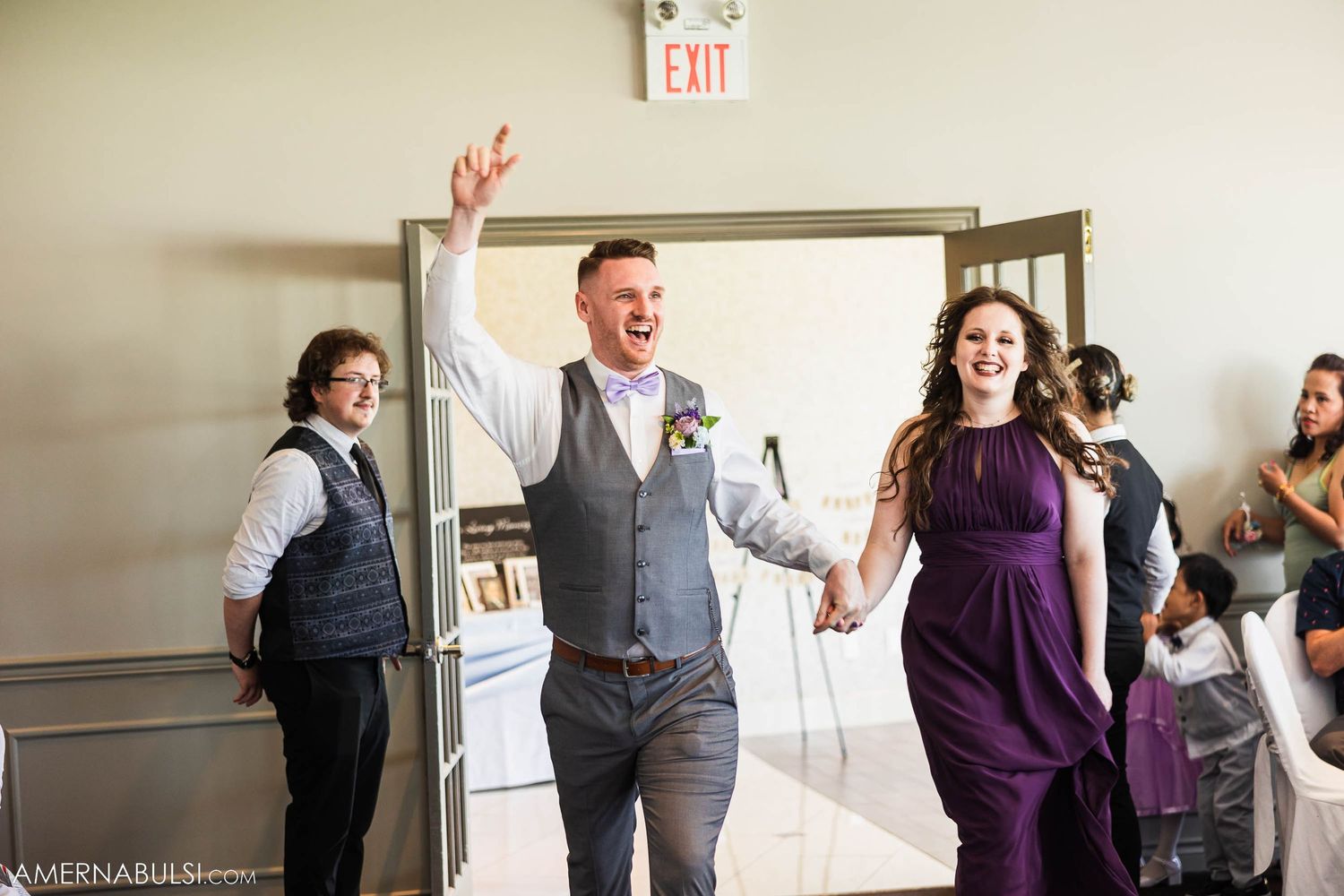 Wedding party members dance excitedly in gray and purple attire at indoor reception.