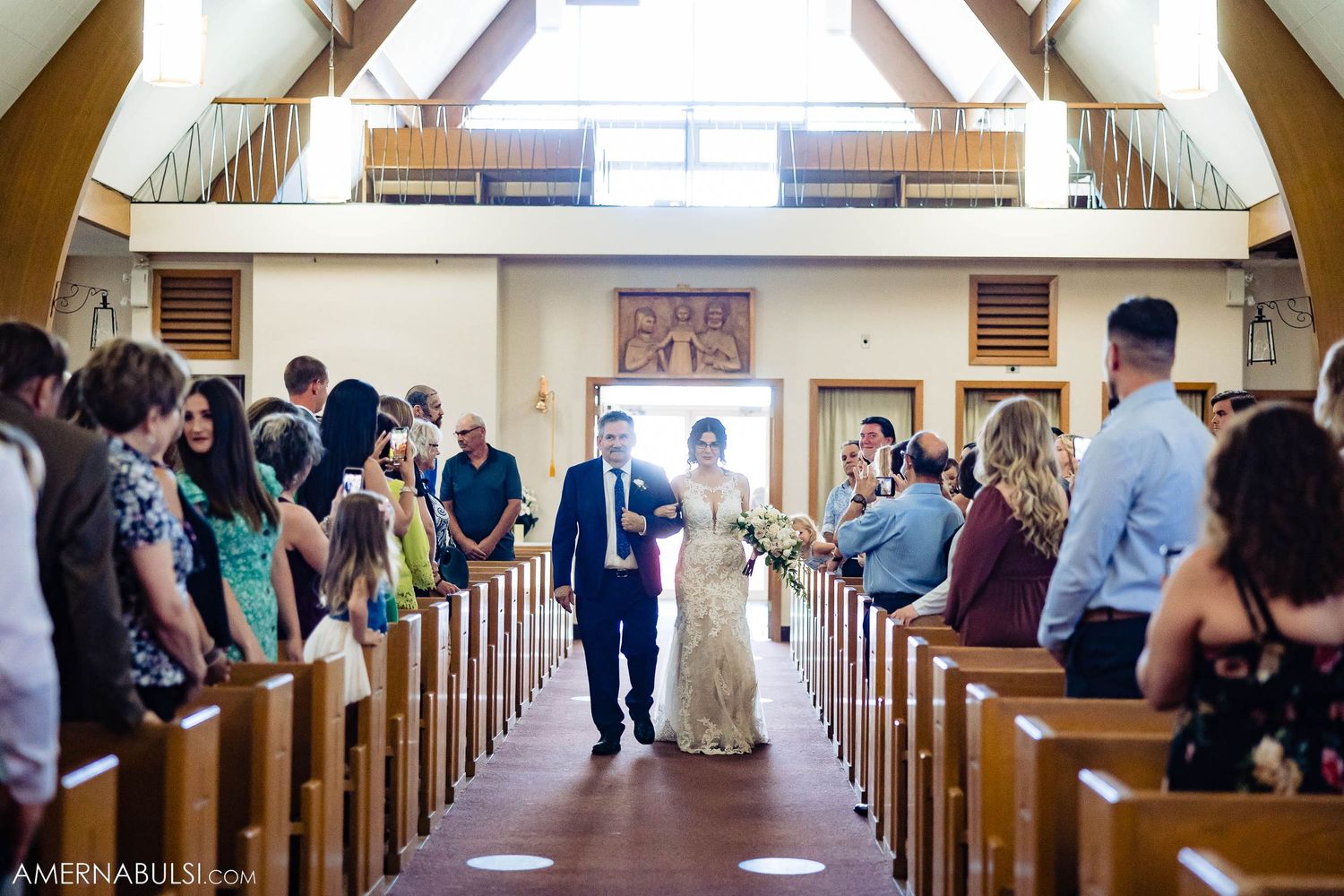 Wedding party members walk down church aisle as guests look on from wooden pews.