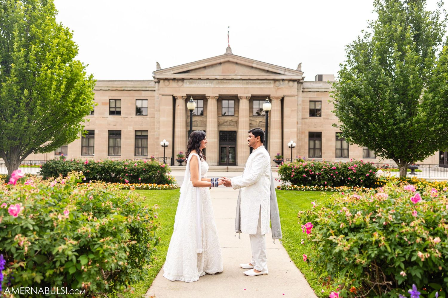Couple standing in front of a neoclassical building surrounded by blooming gardens.