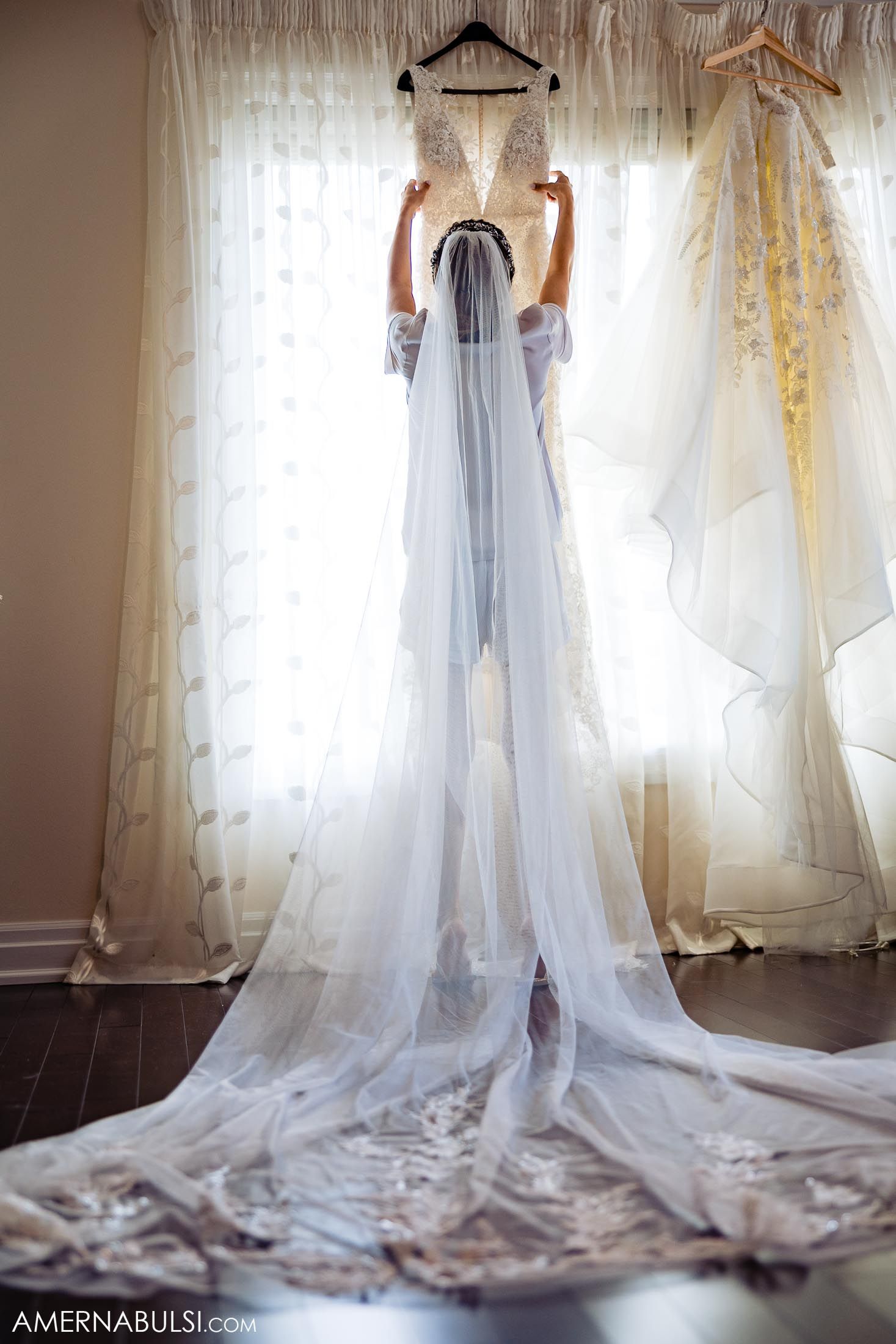 Wedding dress hanging in front of sheer curtains with dramatic train flowing on hardwood floor.