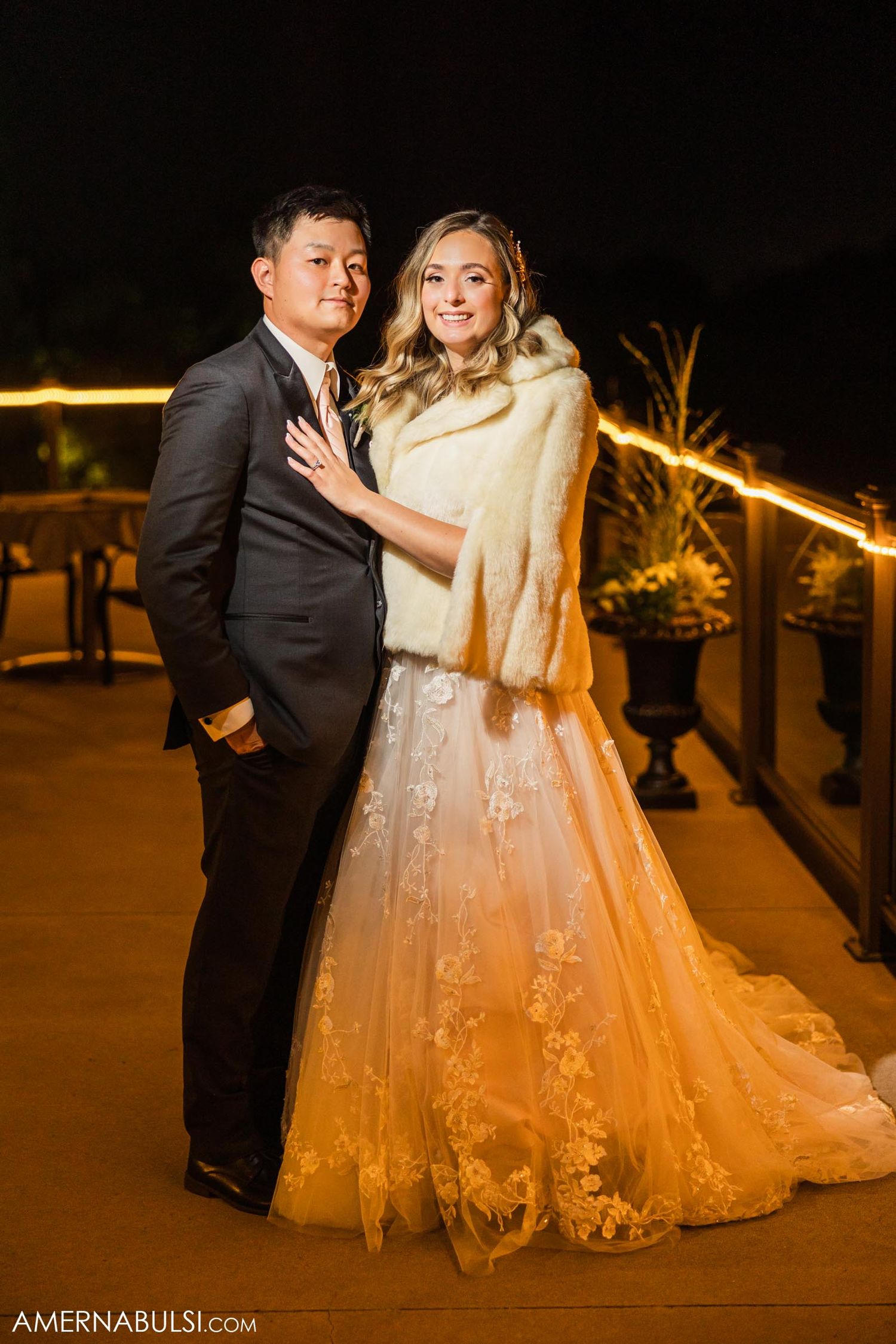 A couple in formal wedding attire poses on a balcony at night.