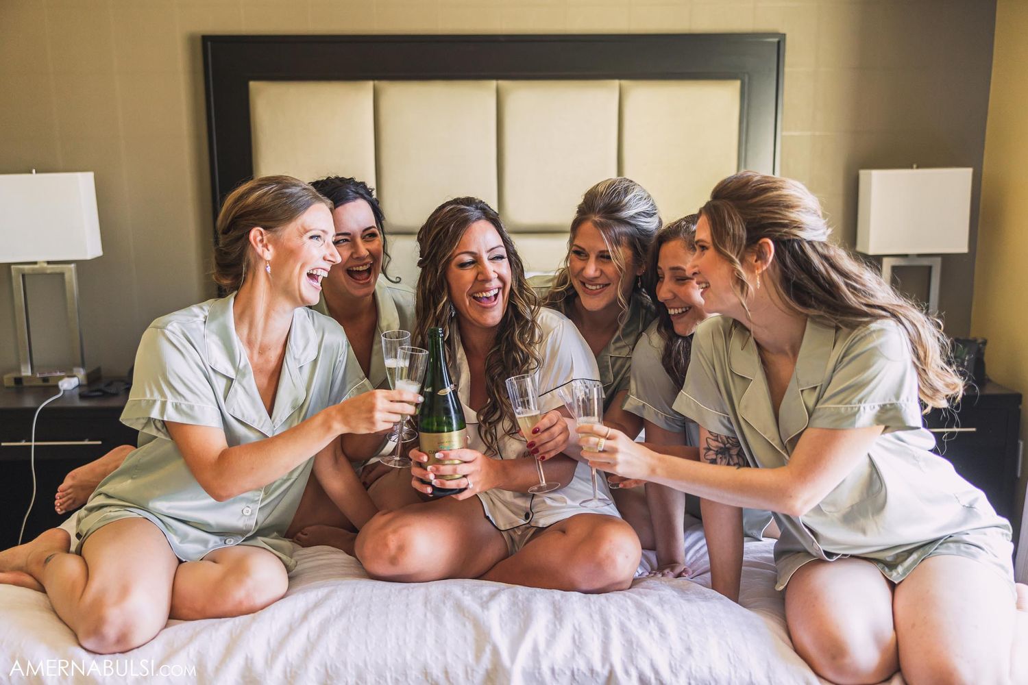 Friends in light colored robes share drinks and laughter while sitting together on a bed.