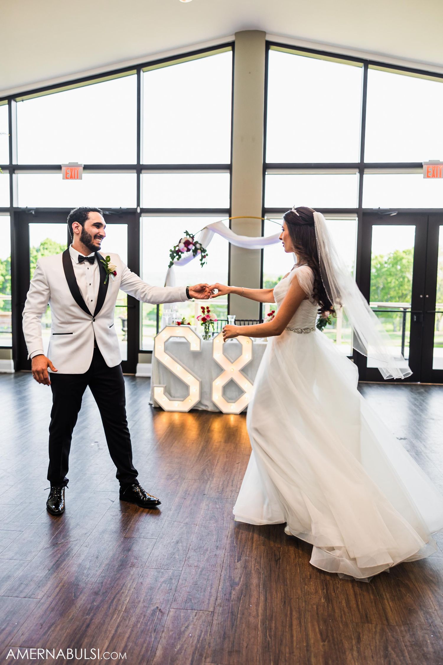 Couple dances in front of illuminated S& letters on wooden floor with floor-to-ceiling windows.