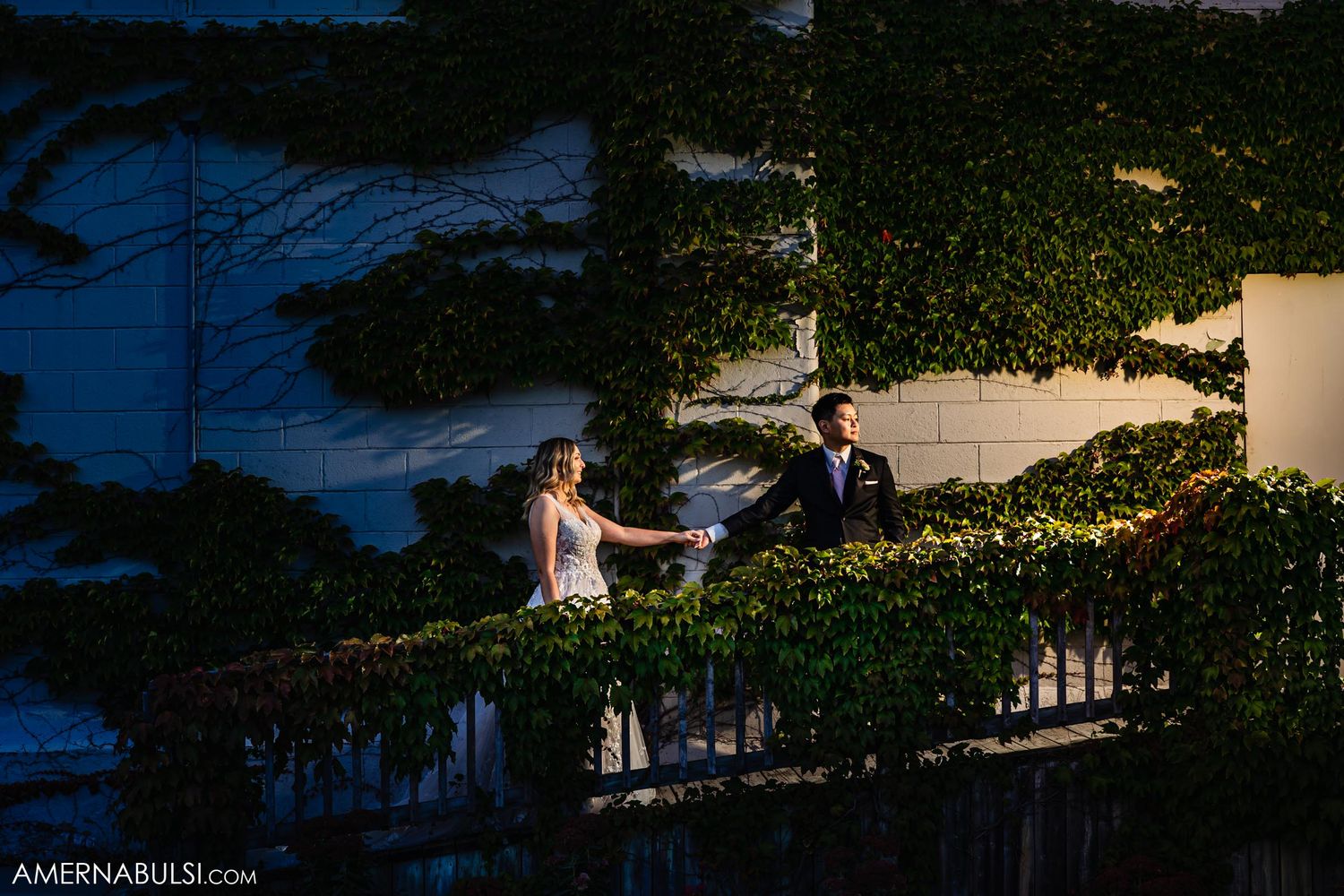 Romantic evening portrait of newlyweds on ivy-covered terrace with dramatic lighting.