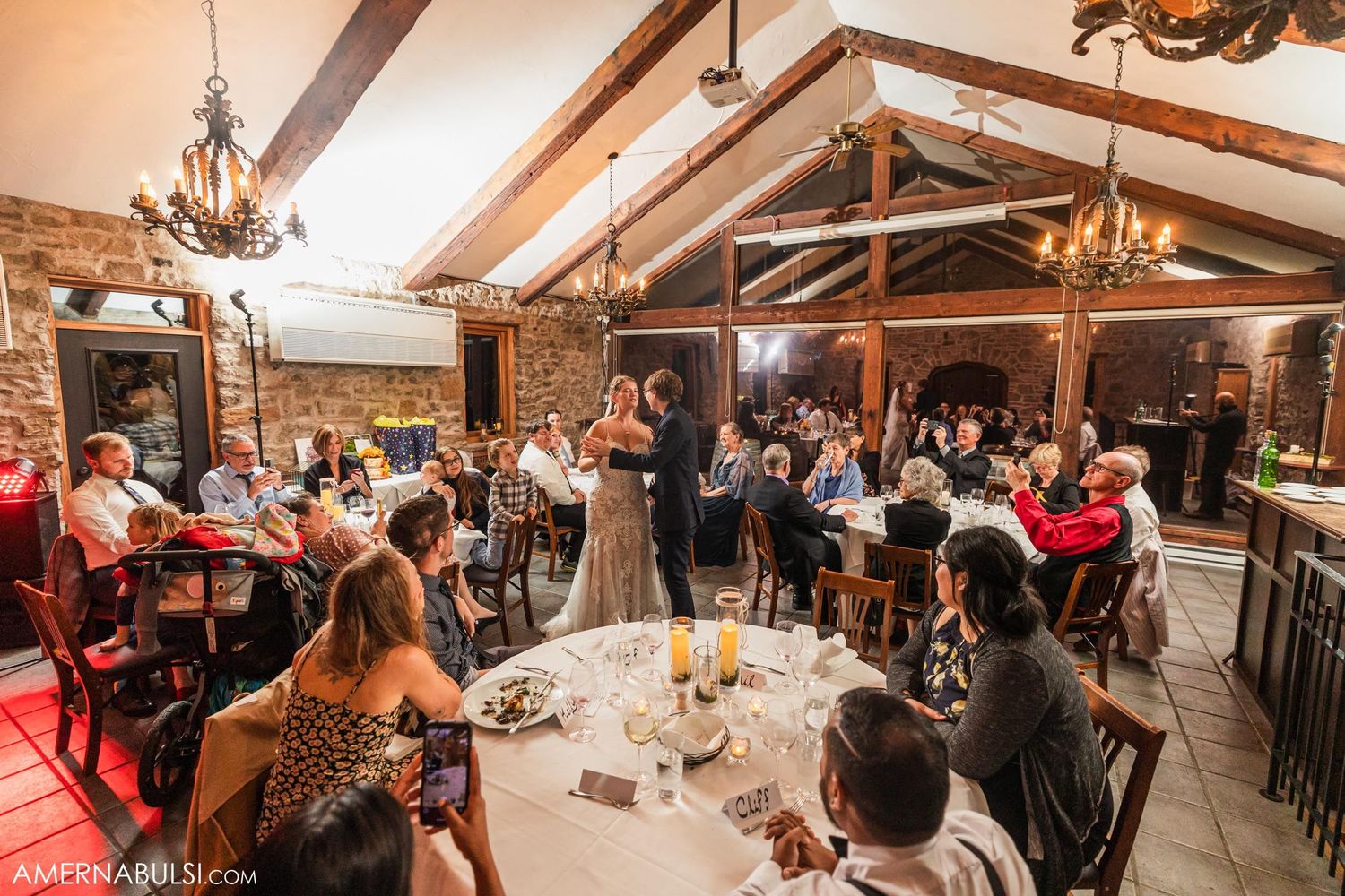 Reception dinner in a rustic venue with vaulted ceilings and chandeliers as guests gather at round tables.