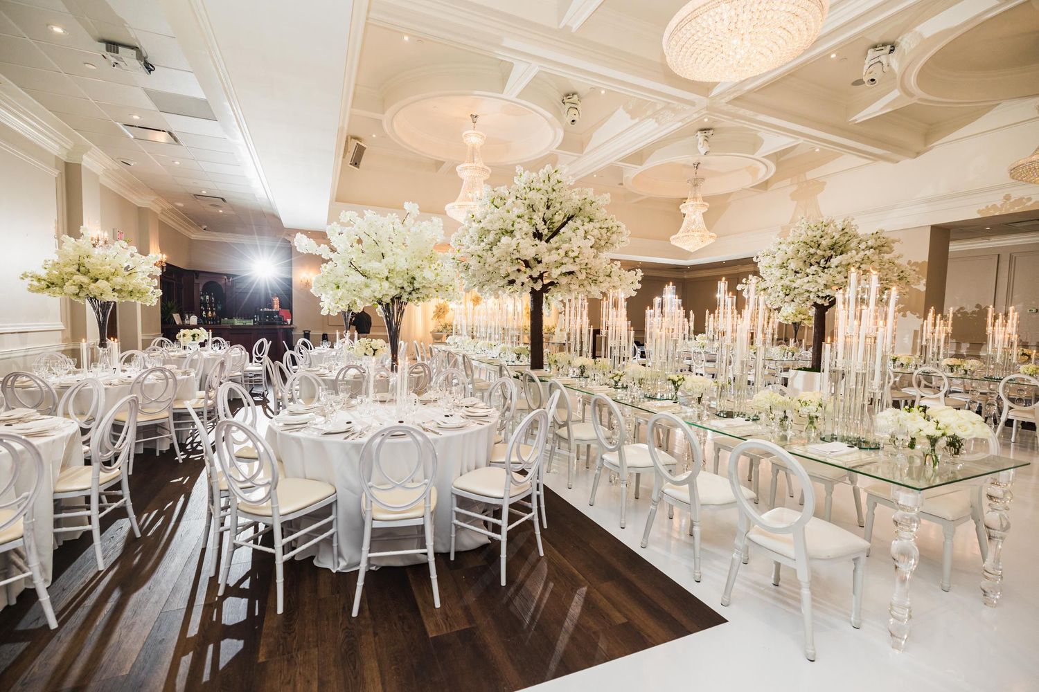 Wedding reception ballroom with tall white floral centerpieces and crystal chandeliers.
