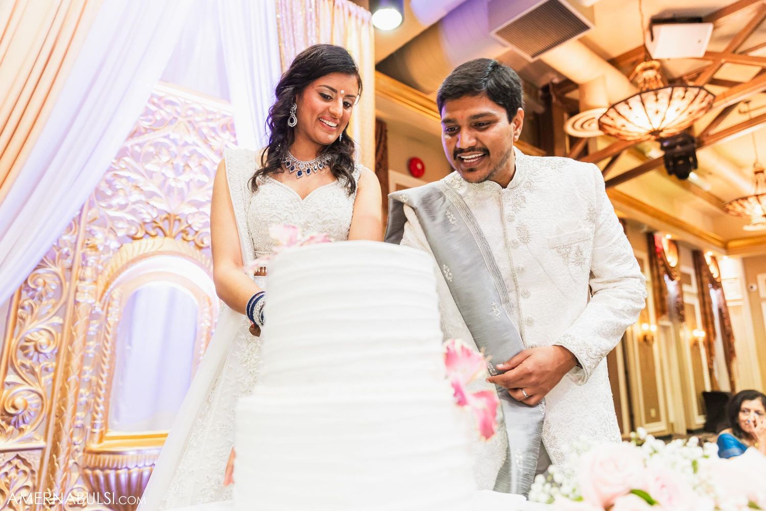 Couple stands together smiling and cutting their elegant white wedding cake.