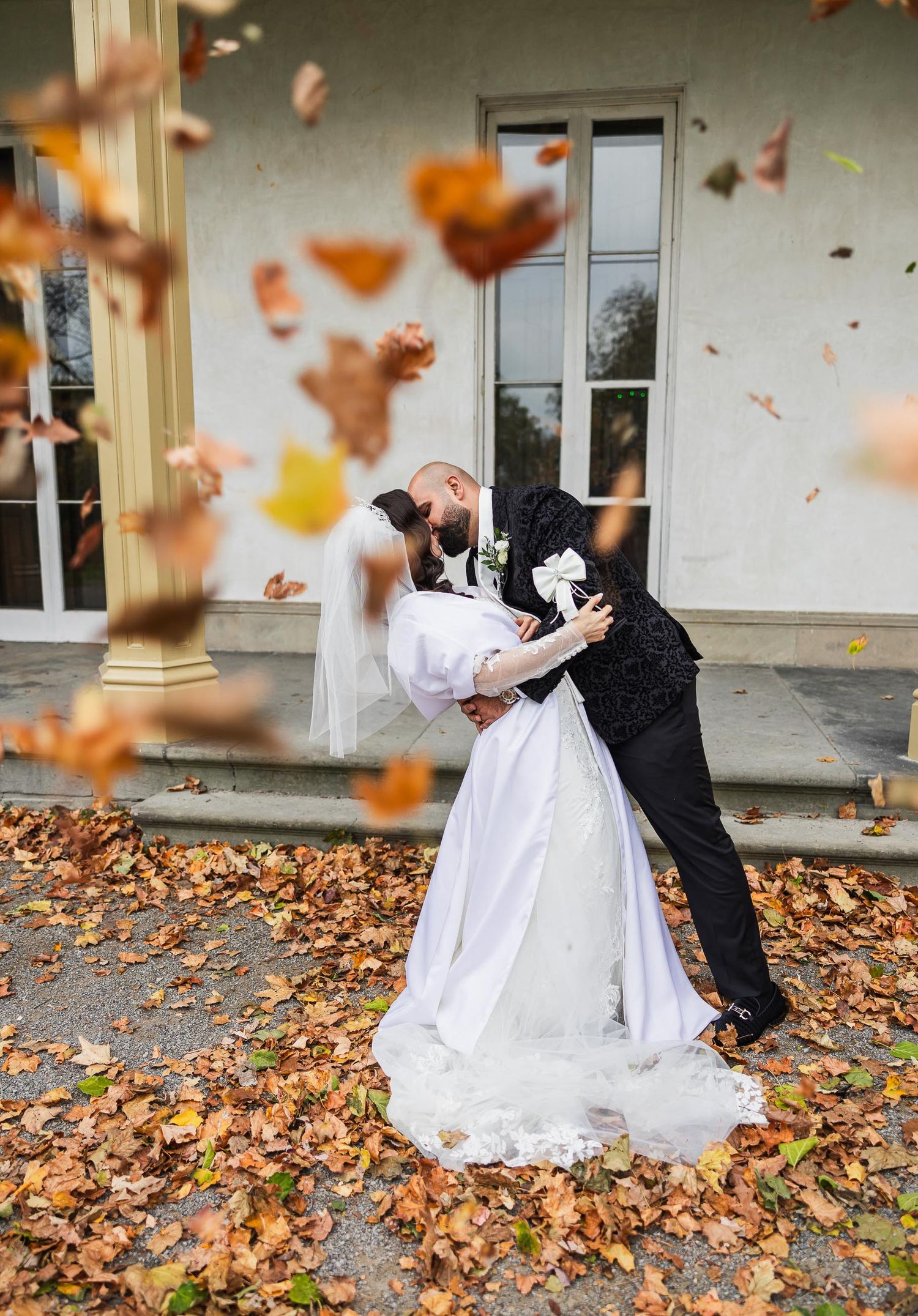 A couple dances surrounded by swirling autumn leaves in front of a classic building.