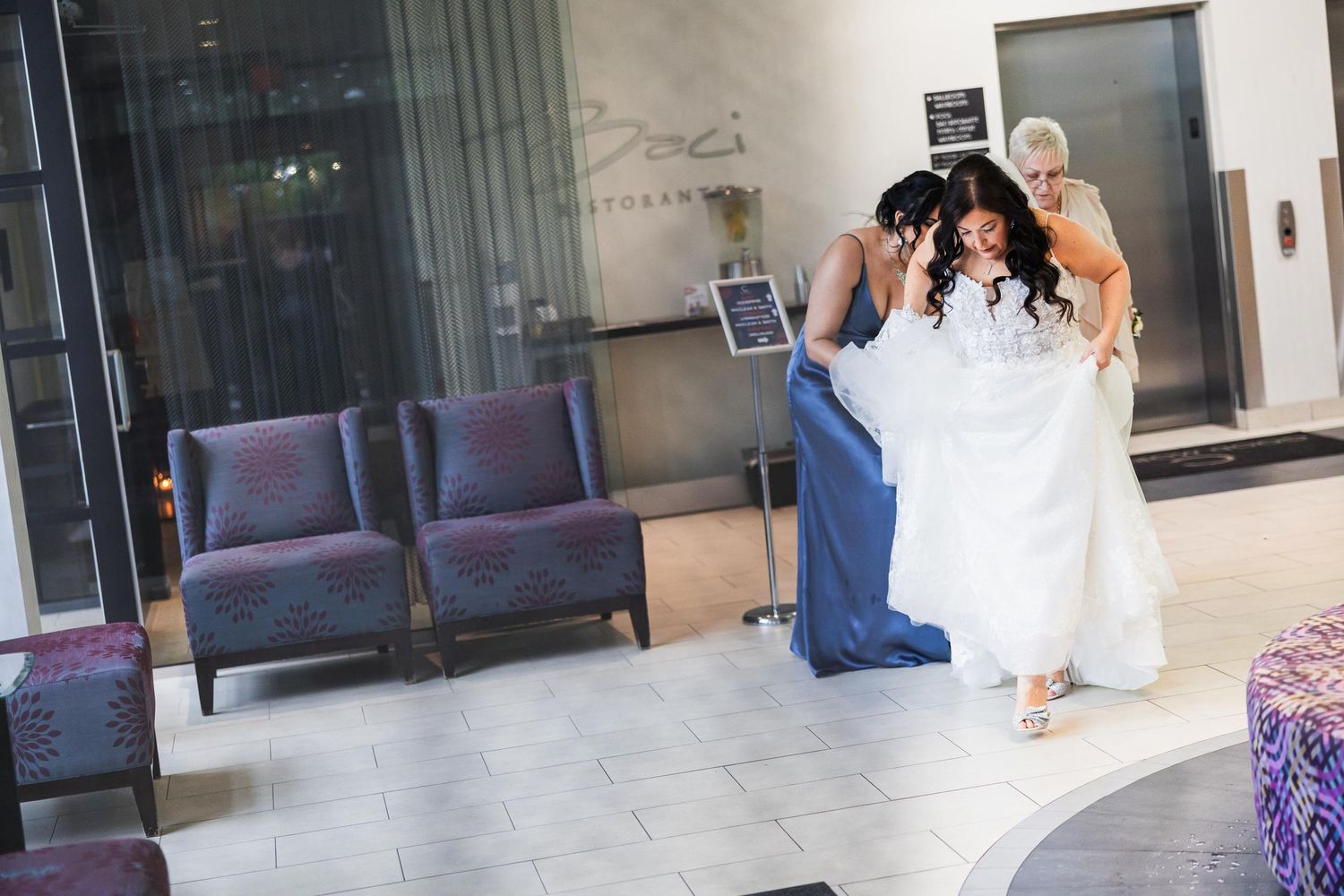 Wedding party helps with final dress adjustments in a hotel lobby with purple chairs.
