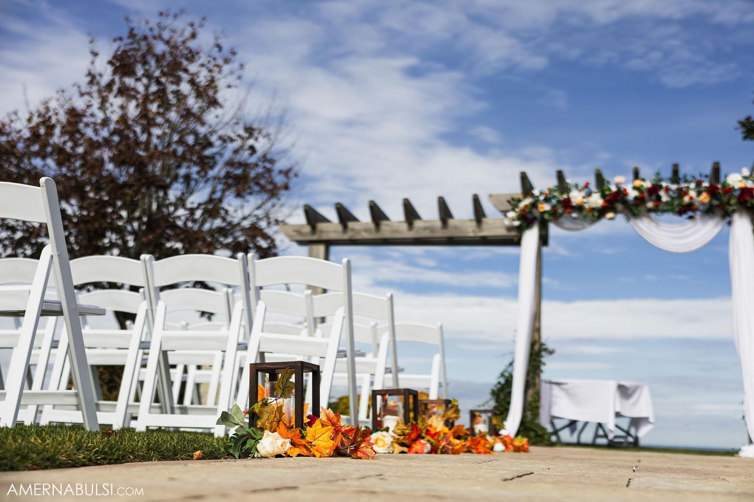 Outdoor wedding ceremony setup with white chairs and floral decorations under a wooden pergola against blue sky.