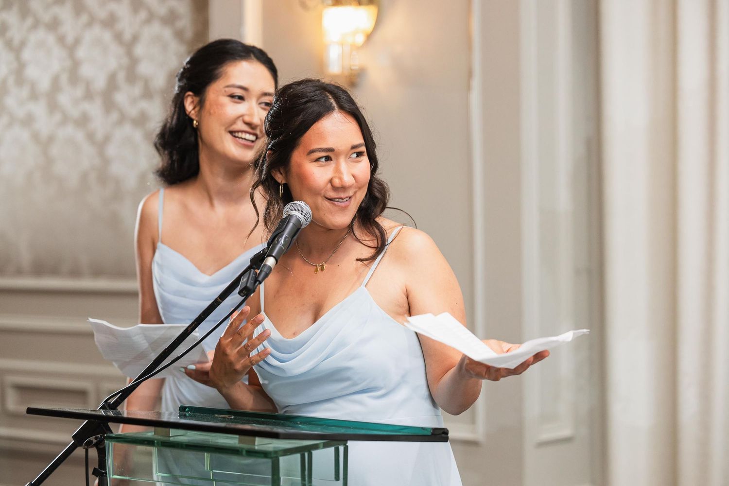 Two bridesmaids in light blue dresses giving joint speech at wedding reception.