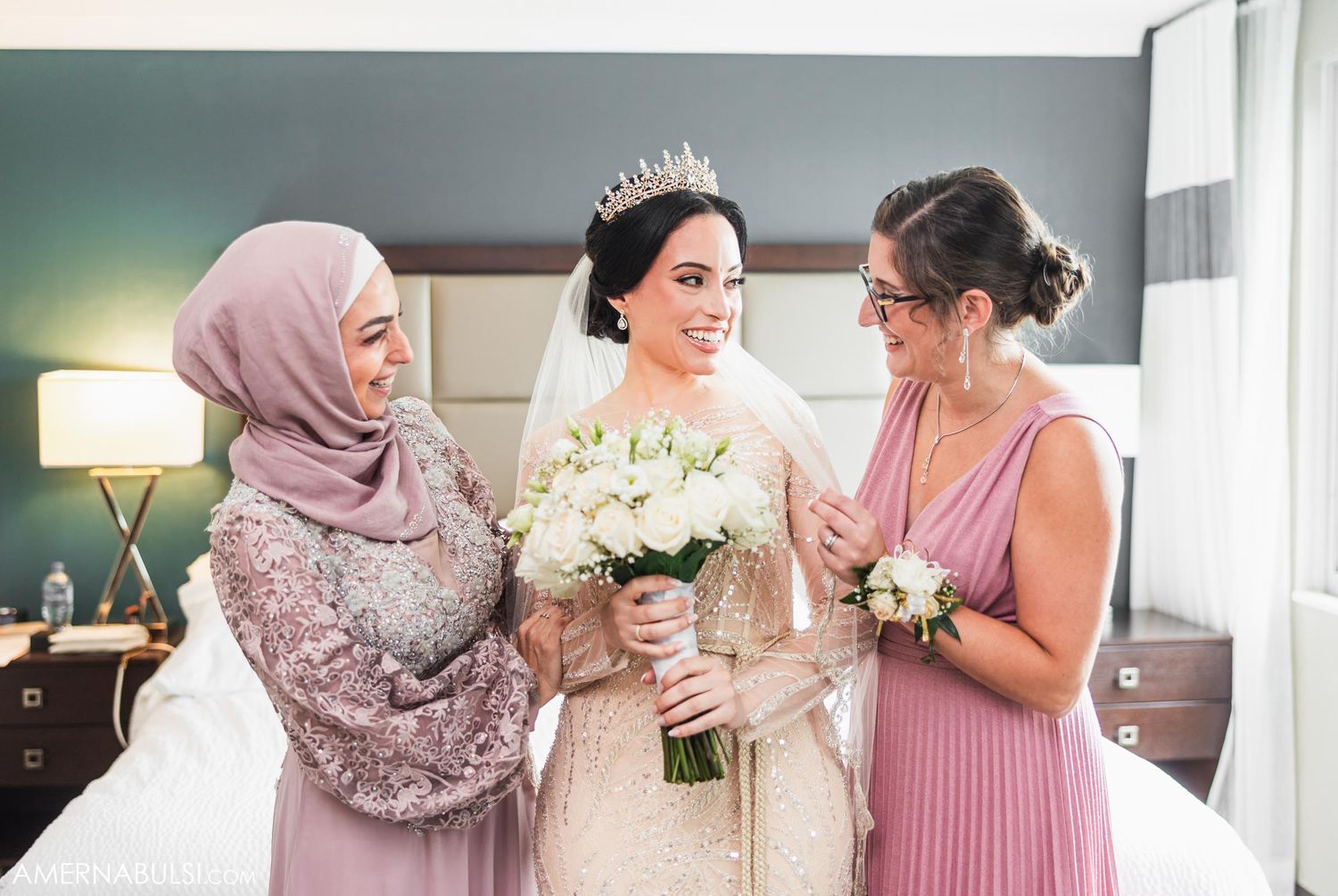 Three women share a tender moment while holding bouquets in elegant wedding attire.