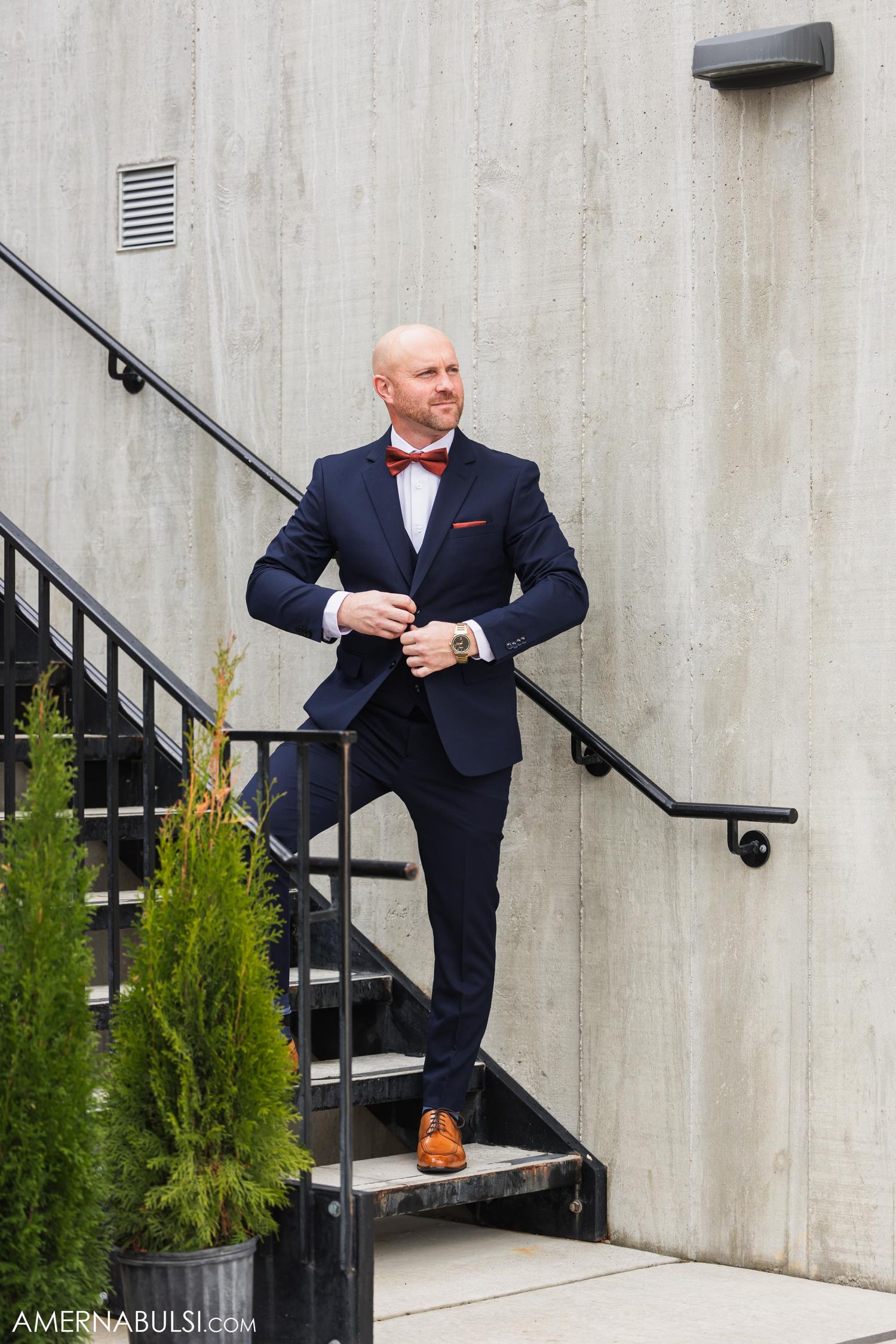 Stylish groom in navy suit with red bowtie poses on concrete staircase.