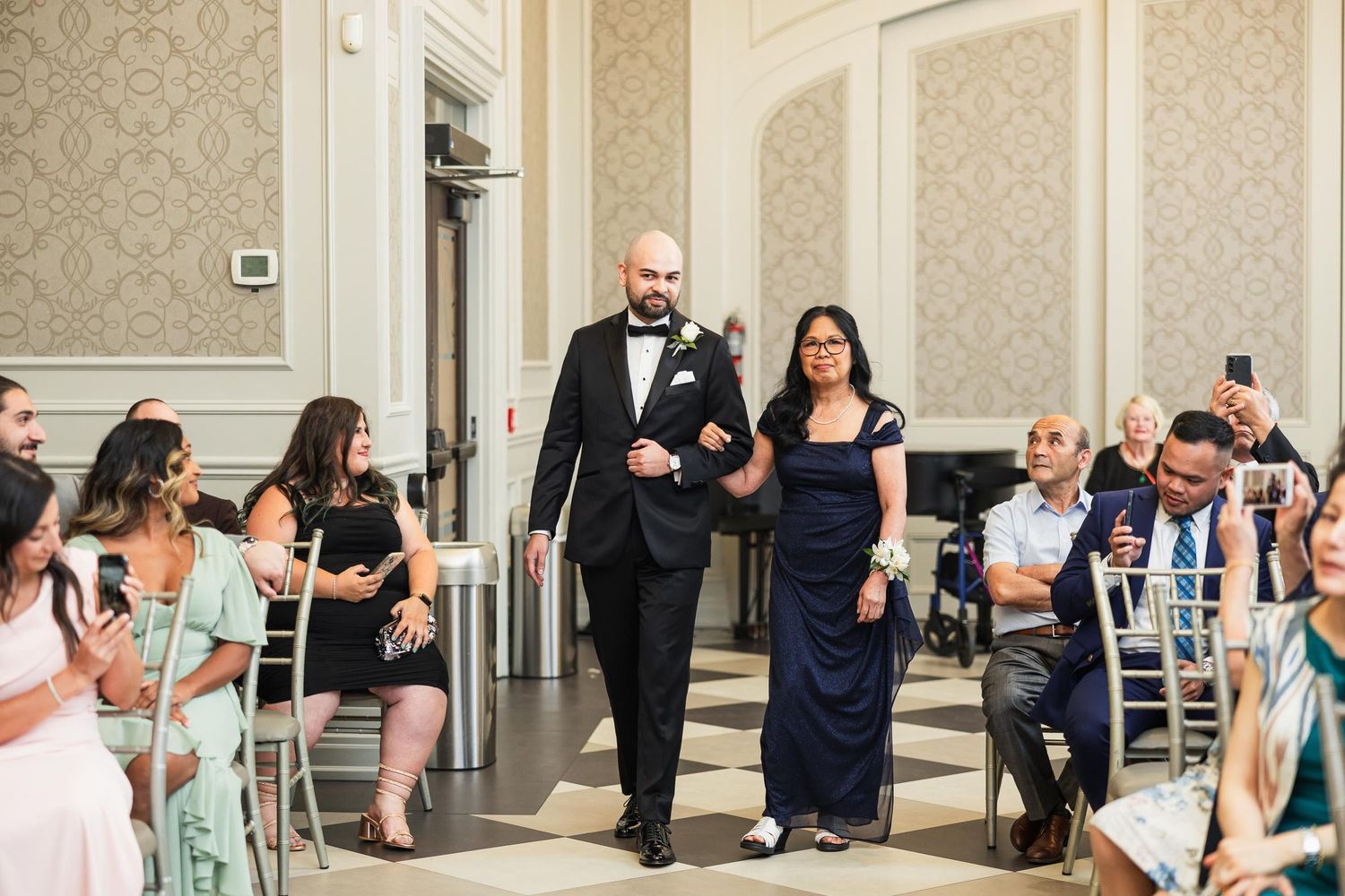 Wedding party members walk down aisle in formal attire as seated guests look on.