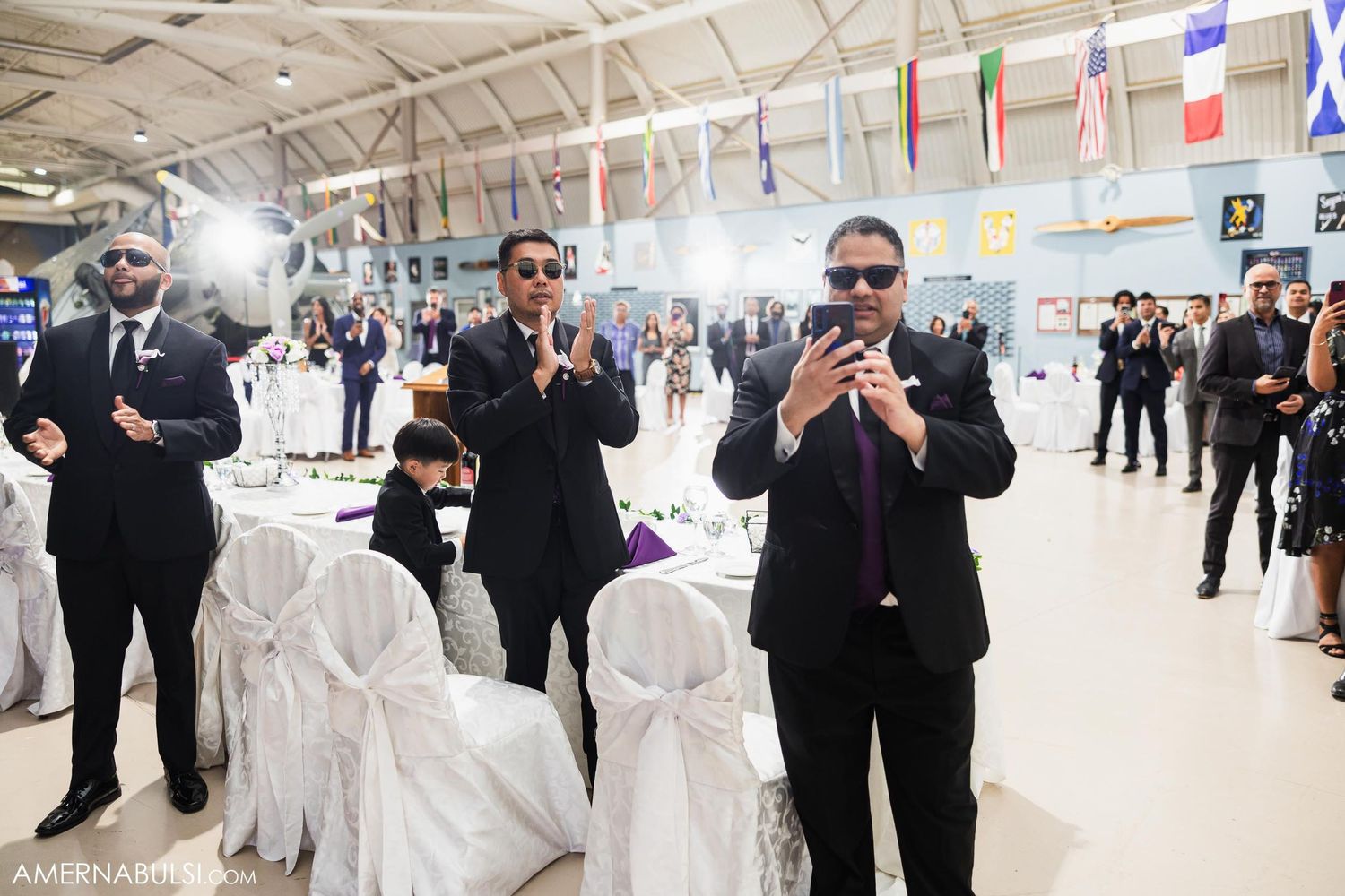 Wedding guests in dark suits celebrate at aviation-themed reception with decorated tables.