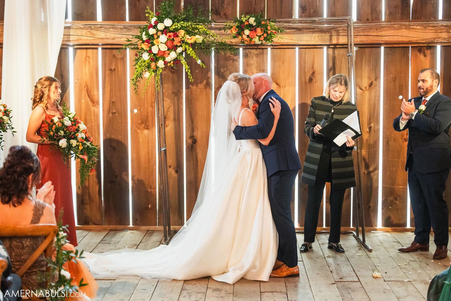 Intimate wedding ceremony moment in barn with floral decorations and wooden backdrop.