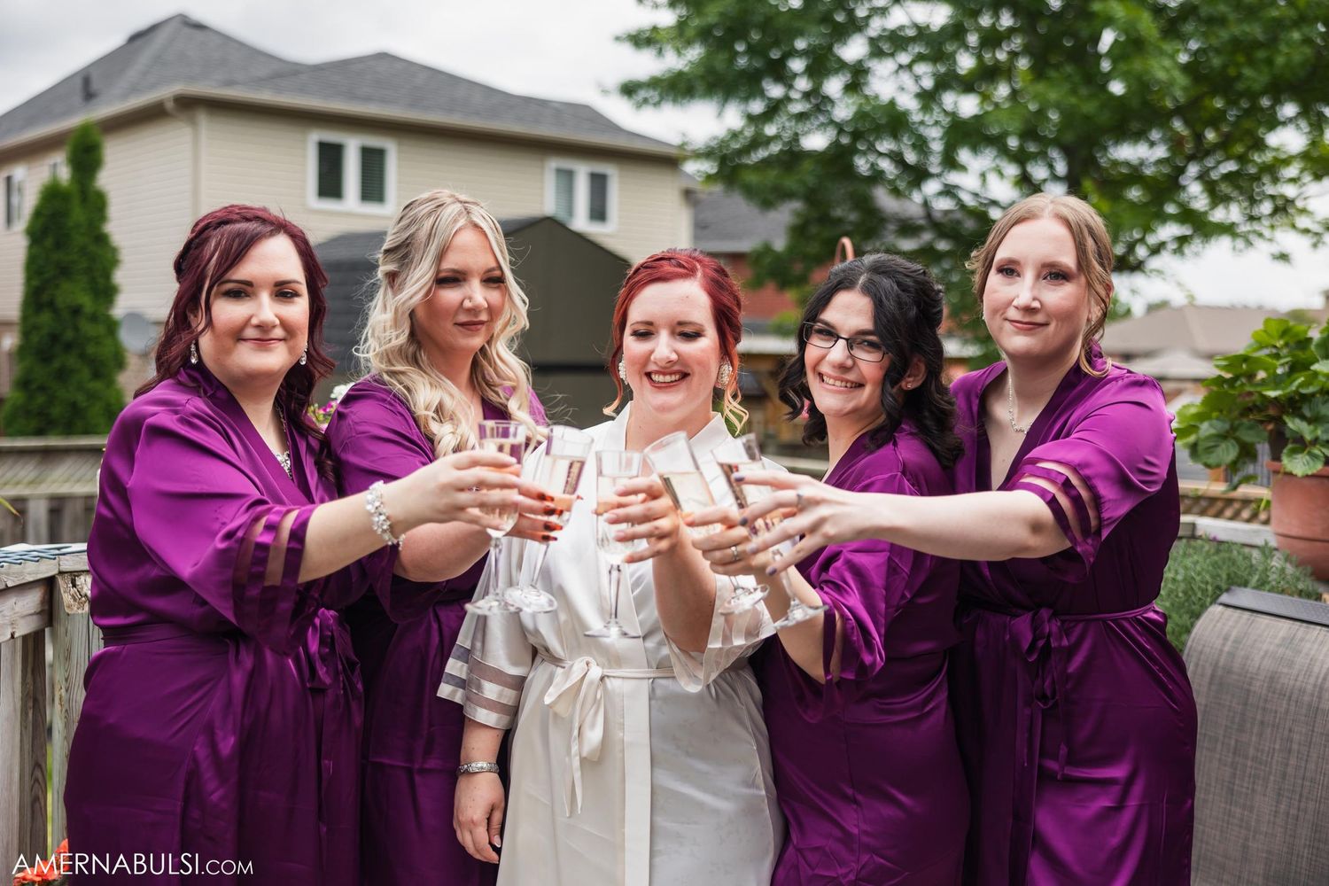 Bridal party in purple robes toasting champagne glasses on outdoor deck.