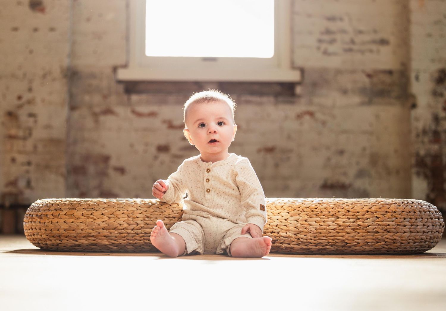 A baby in cream-colored clothing sits on a woven mat near a bright window.