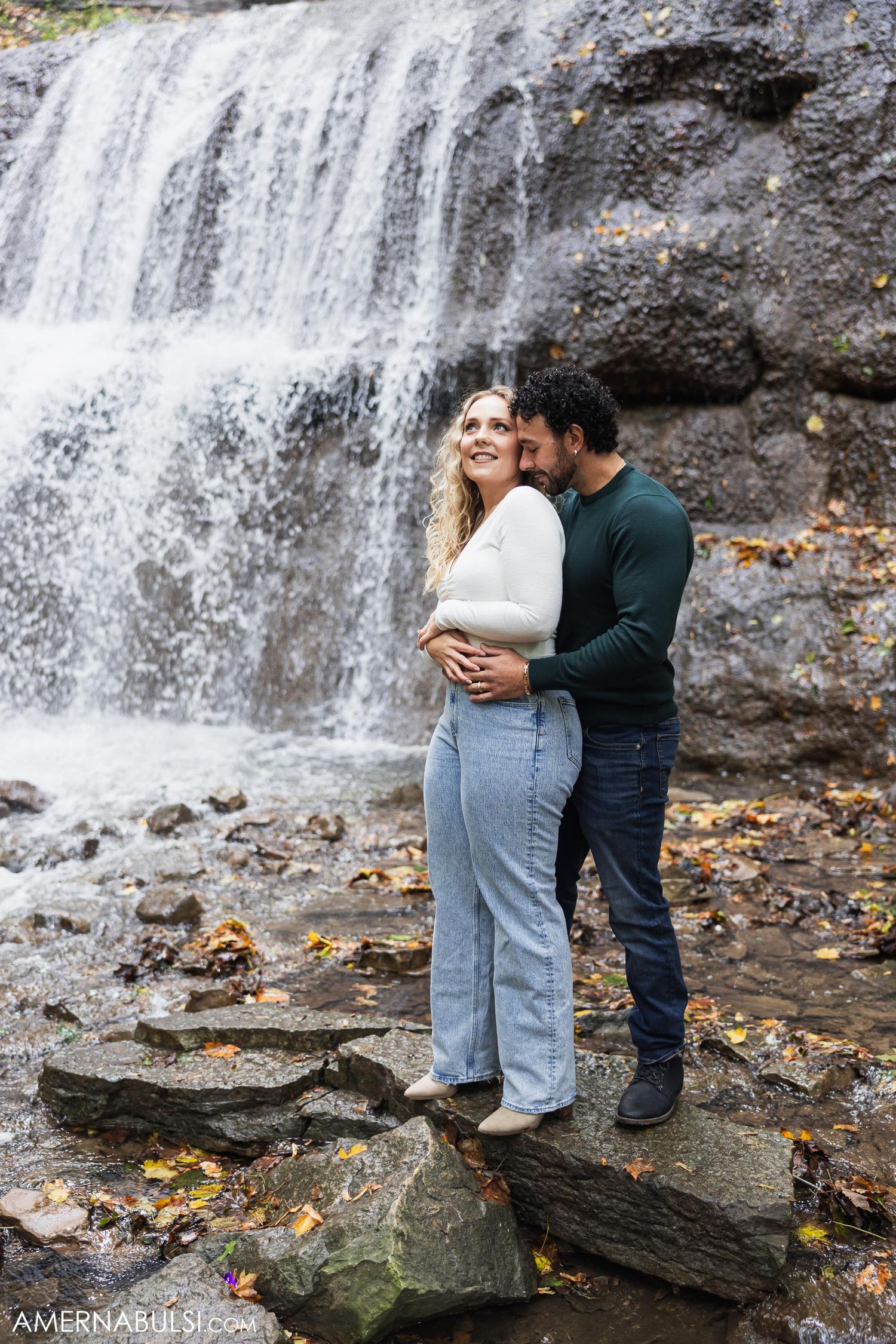 A romantic couple embraces in front of a cascading waterfall surrounded by autumn leaves and rocky terrain.