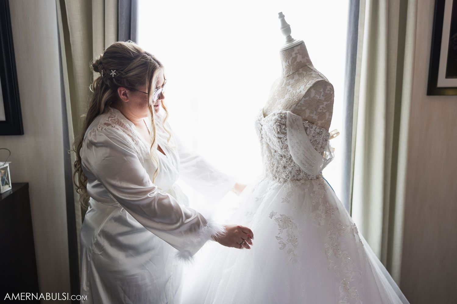 Final adjustments being made to an ornate white wedding dress on a mannequin form near a window.