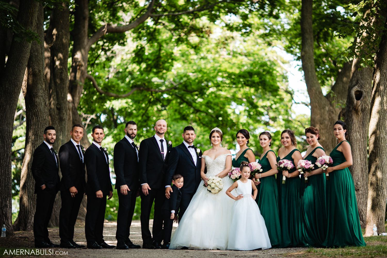 Wedding party lined up outdoors under green trees for formal group portrait in summer.