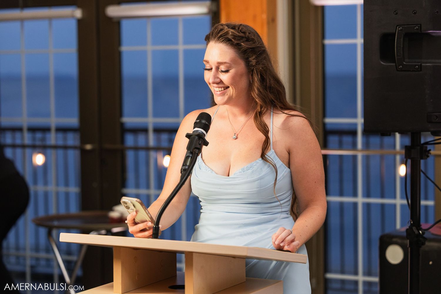 Speaker delivers emotional wedding toast at elegant reception with waterfront backdrop through windows.