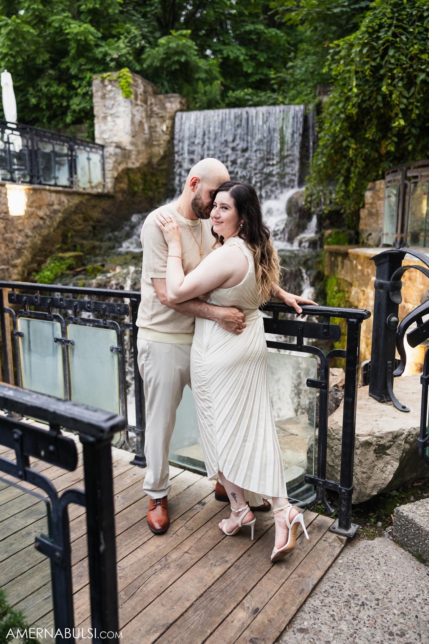 Elegant couple embracing on a wooden deck with a scenic waterfall backdrop.