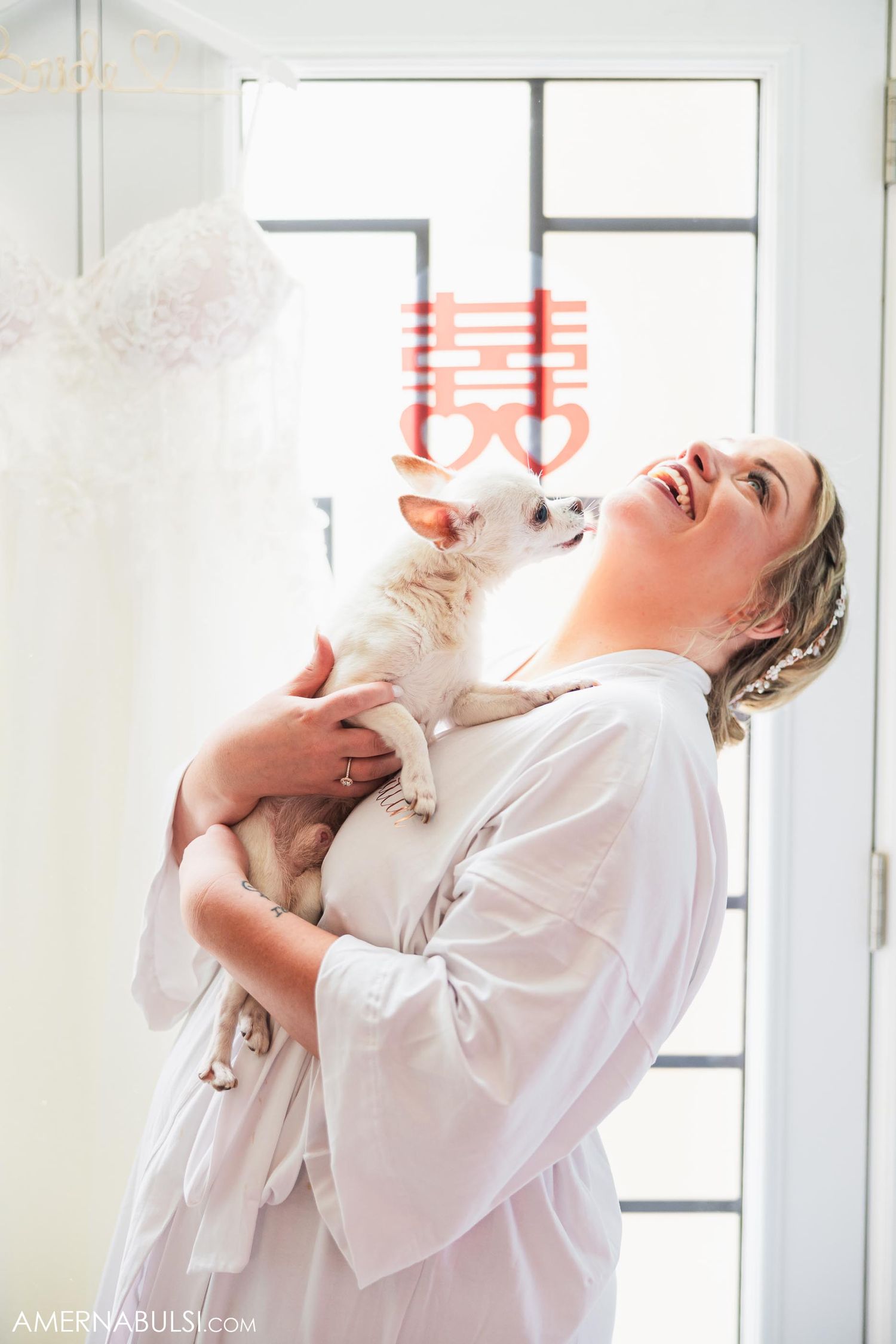Person in white robe laughing while holding small white dog in front of window with Chinese double happiness symbol.