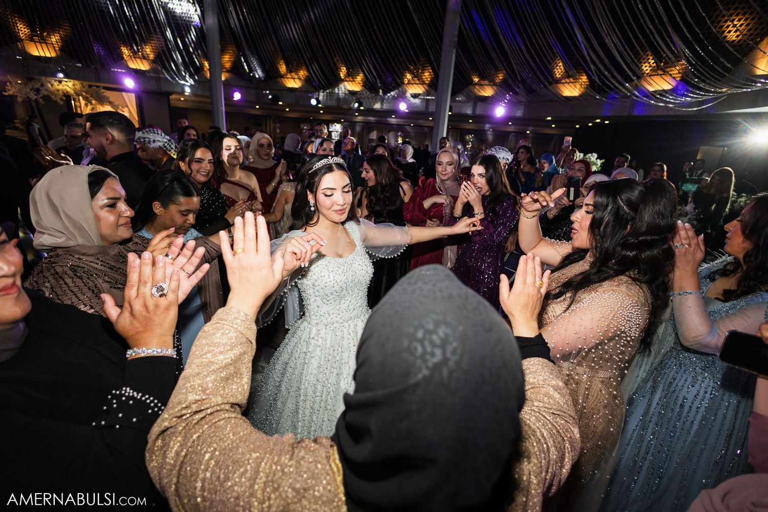 Wedding reception crowd dancing and celebrating with raised hands under colorful lighting at a lively party.