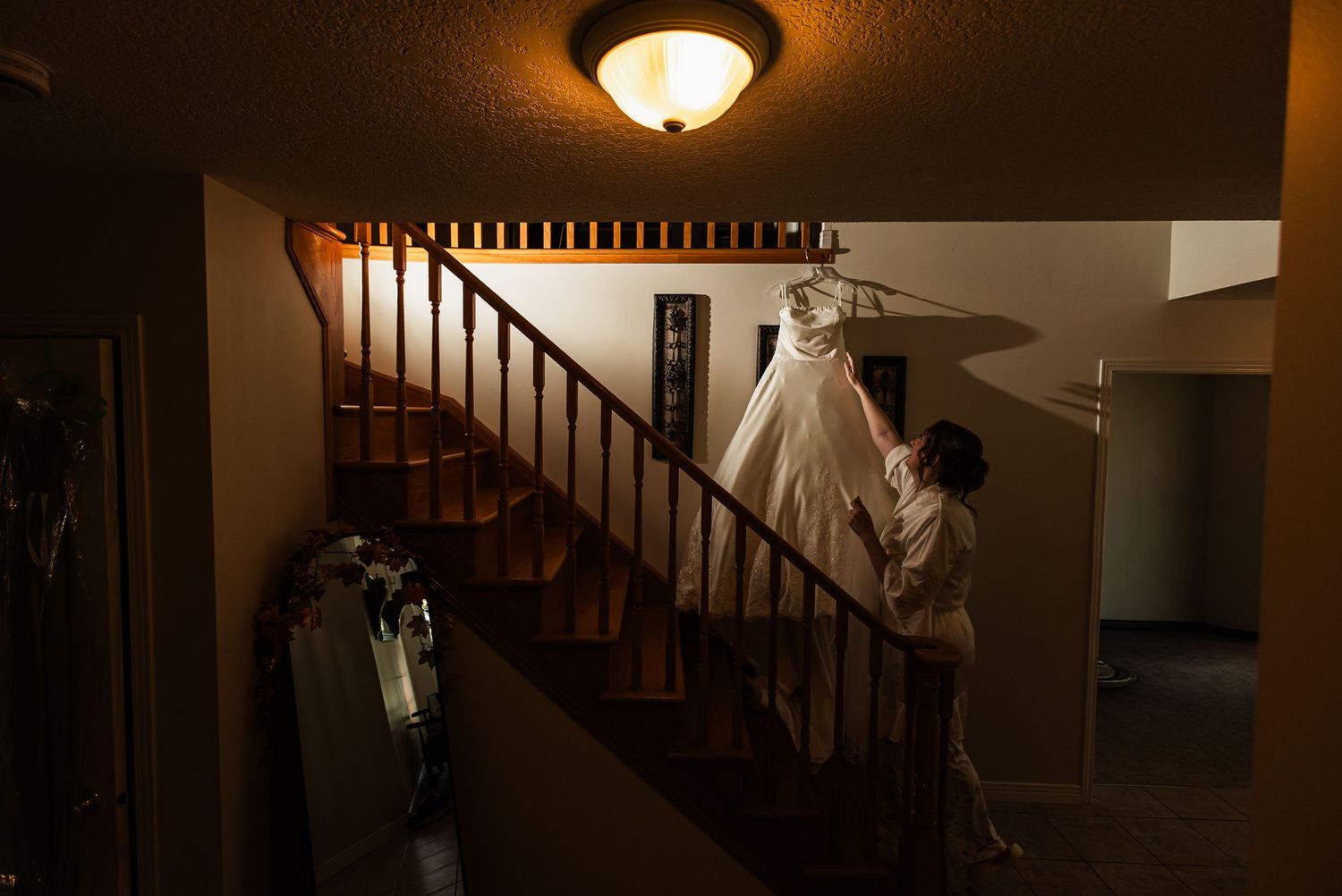 Bride admiring wedding dress hanging on staircase railing in warmly lit home interior.