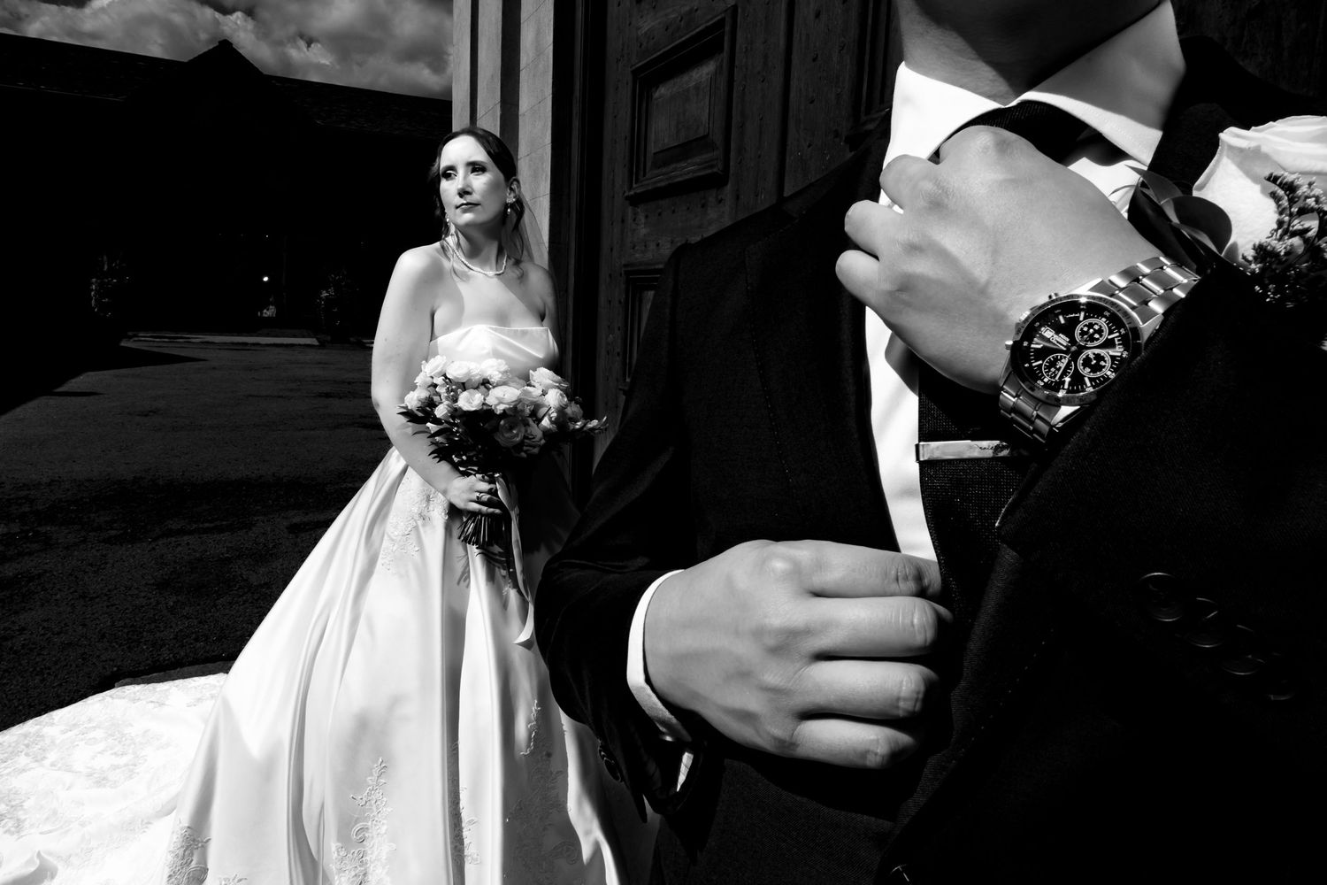Black and white photo of bride holding bouquet standing behind groom adjusting his suit cufflinks.