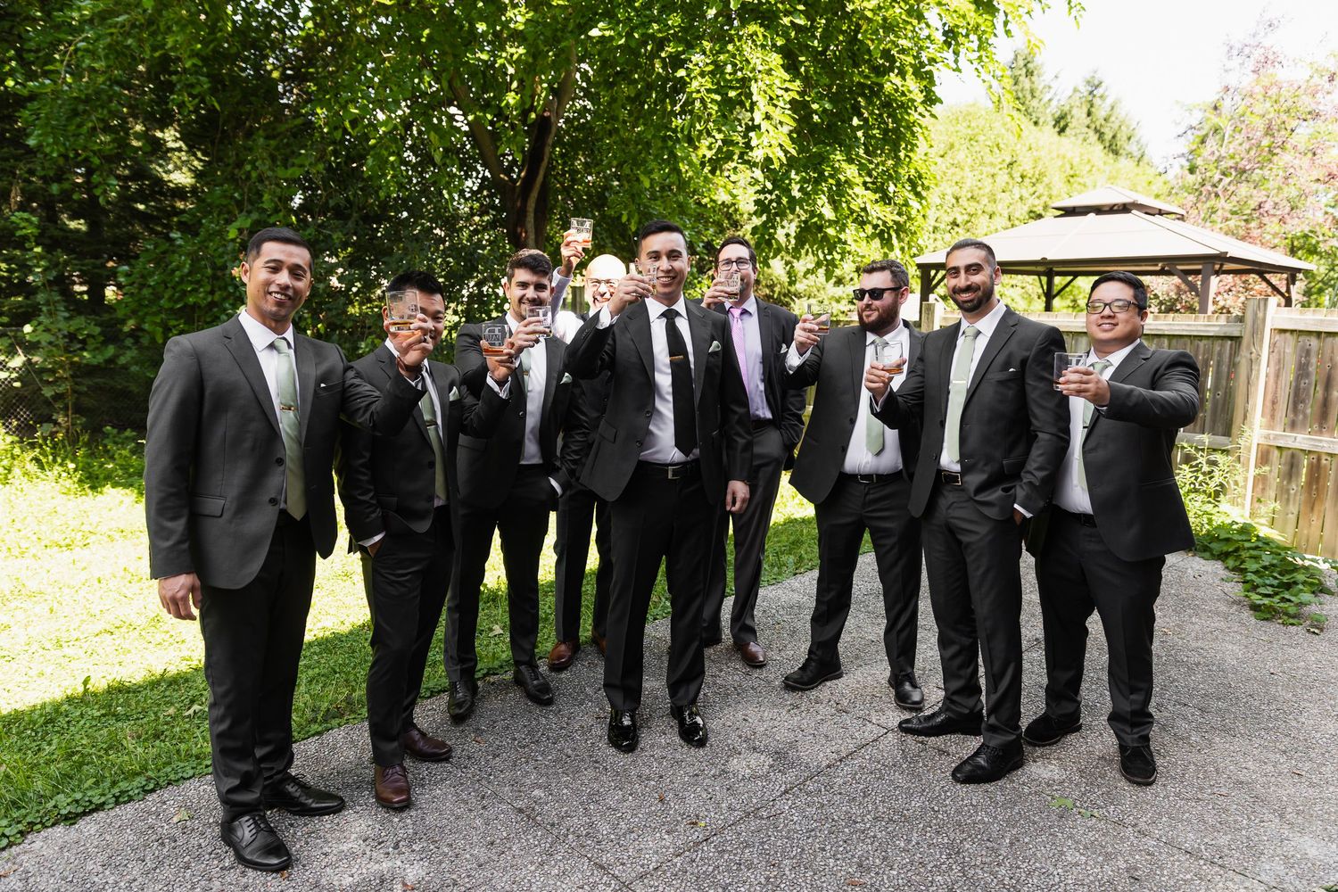 Groomsmen in black suits toast outdoors with drinks in lush garden setting at wedding celebration.
