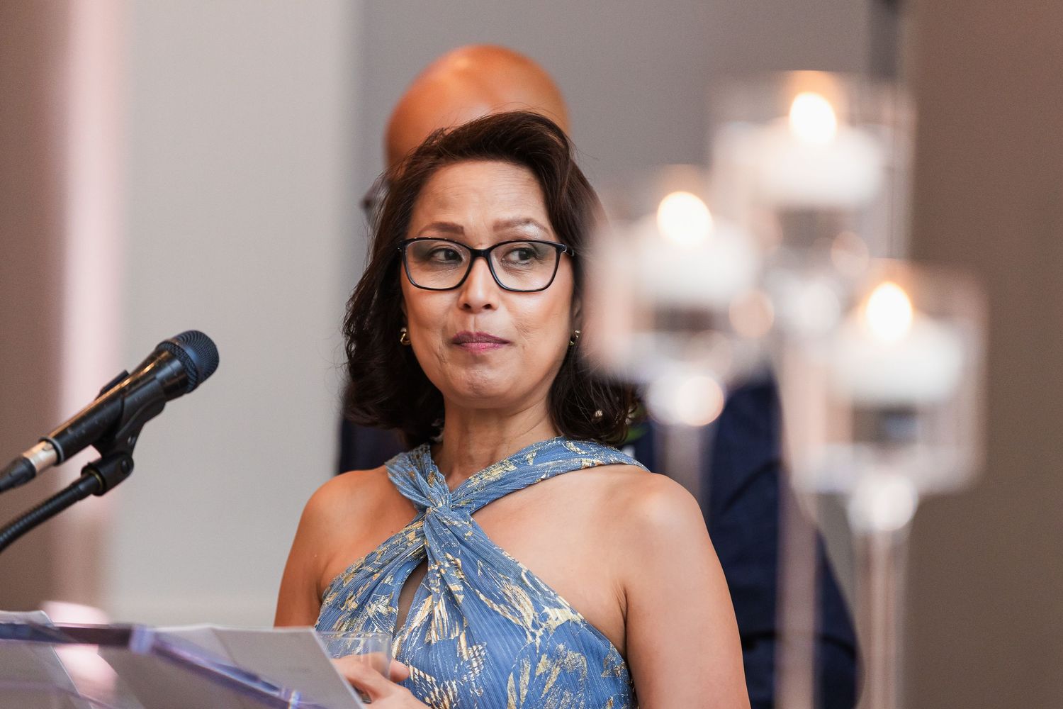 Woman in blue floral dress speaking at podium with candles in background at formal event.