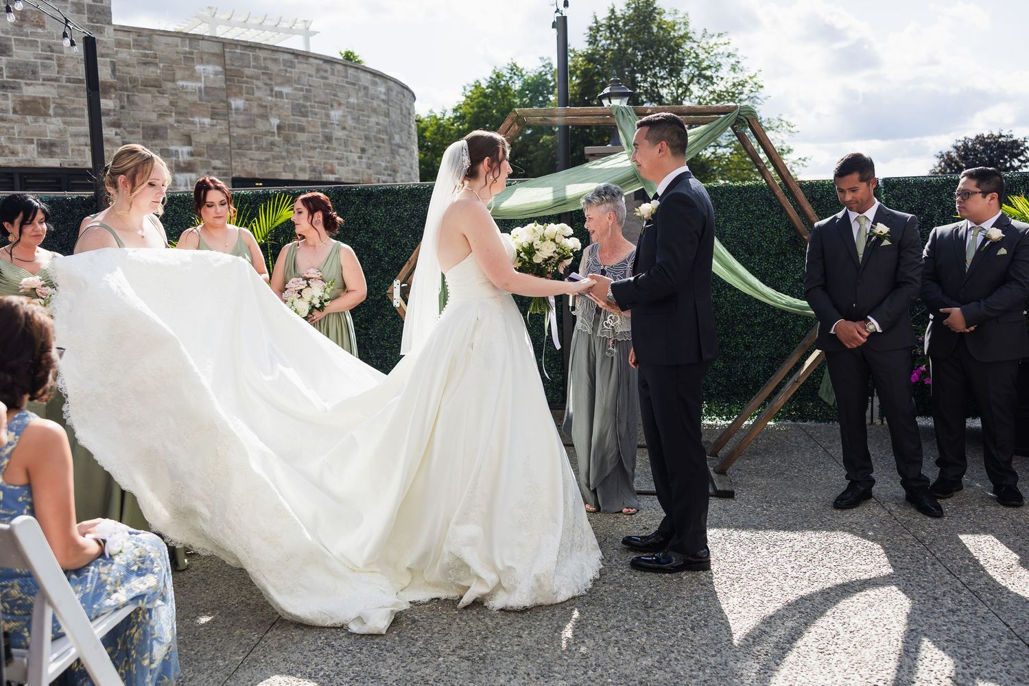 Bride in flowing white gown exchanges vows with groom surrounded by bridesmaids and groomsmen outdoors.
