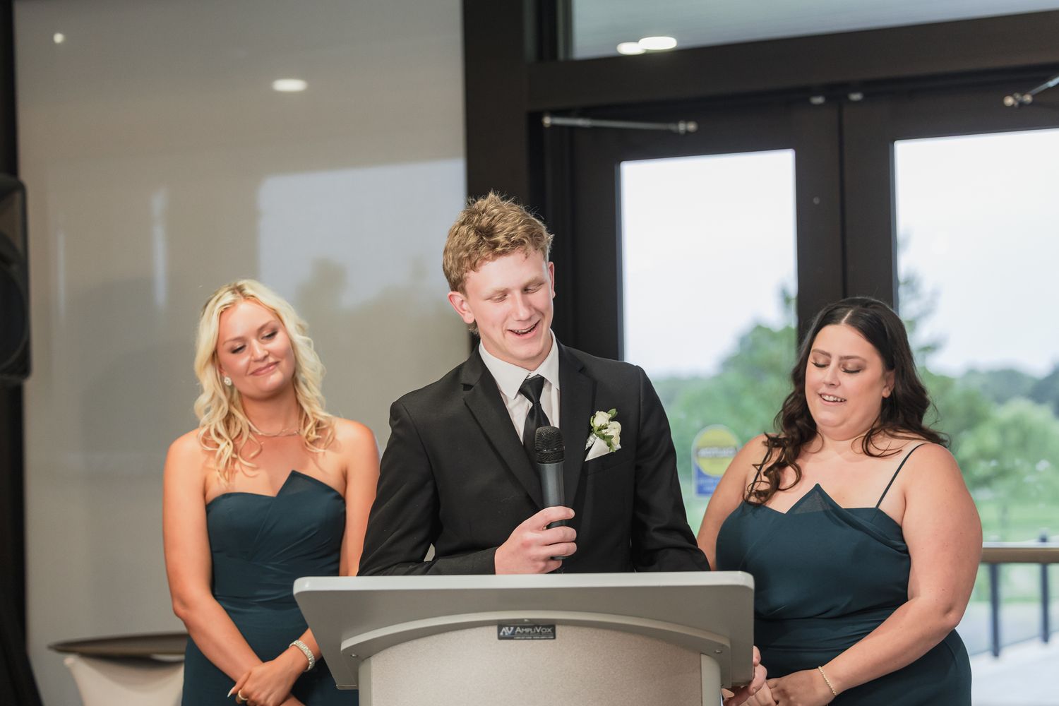 Young man in black tuxedo delivers a wedding speech at podium flanked by two bridesmaids in teal gowns.