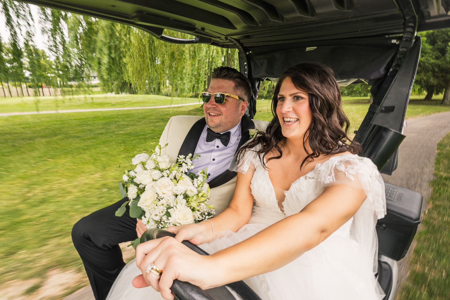 Bride and groom ride golf cart outdoors on wedding day, bride holding white floral bouquet.