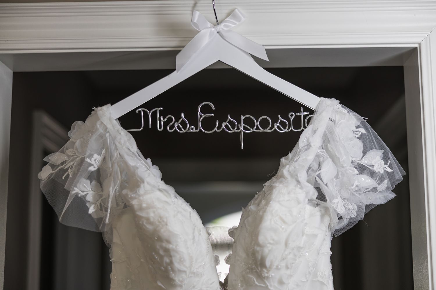 A lace wedding dress hangs on a custom wire hanger reading Mrs. Esposito on a white door frame.