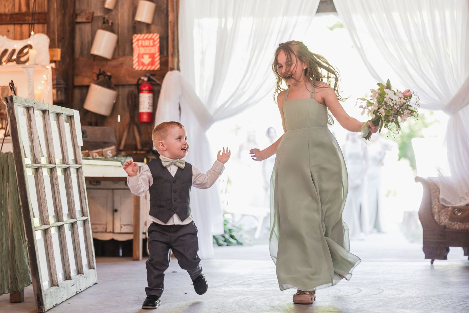 A young ring bearer runs joyfully toward a bridesmaid in a sage green gown holding a bouquet inside a rustic barn wedding venue.