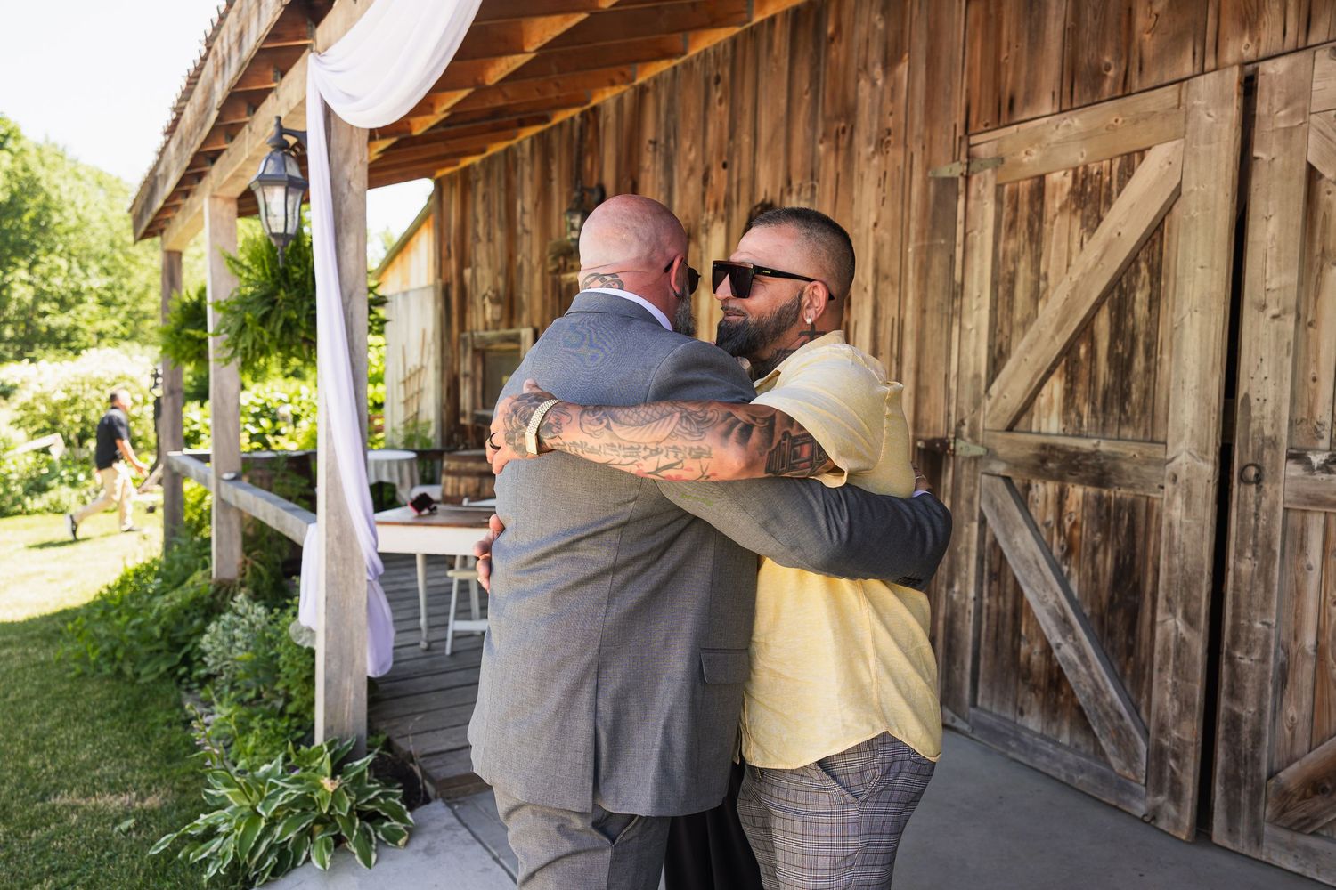 Two men share a warm embrace outside a rustic barn wedding venue on a sunny day.