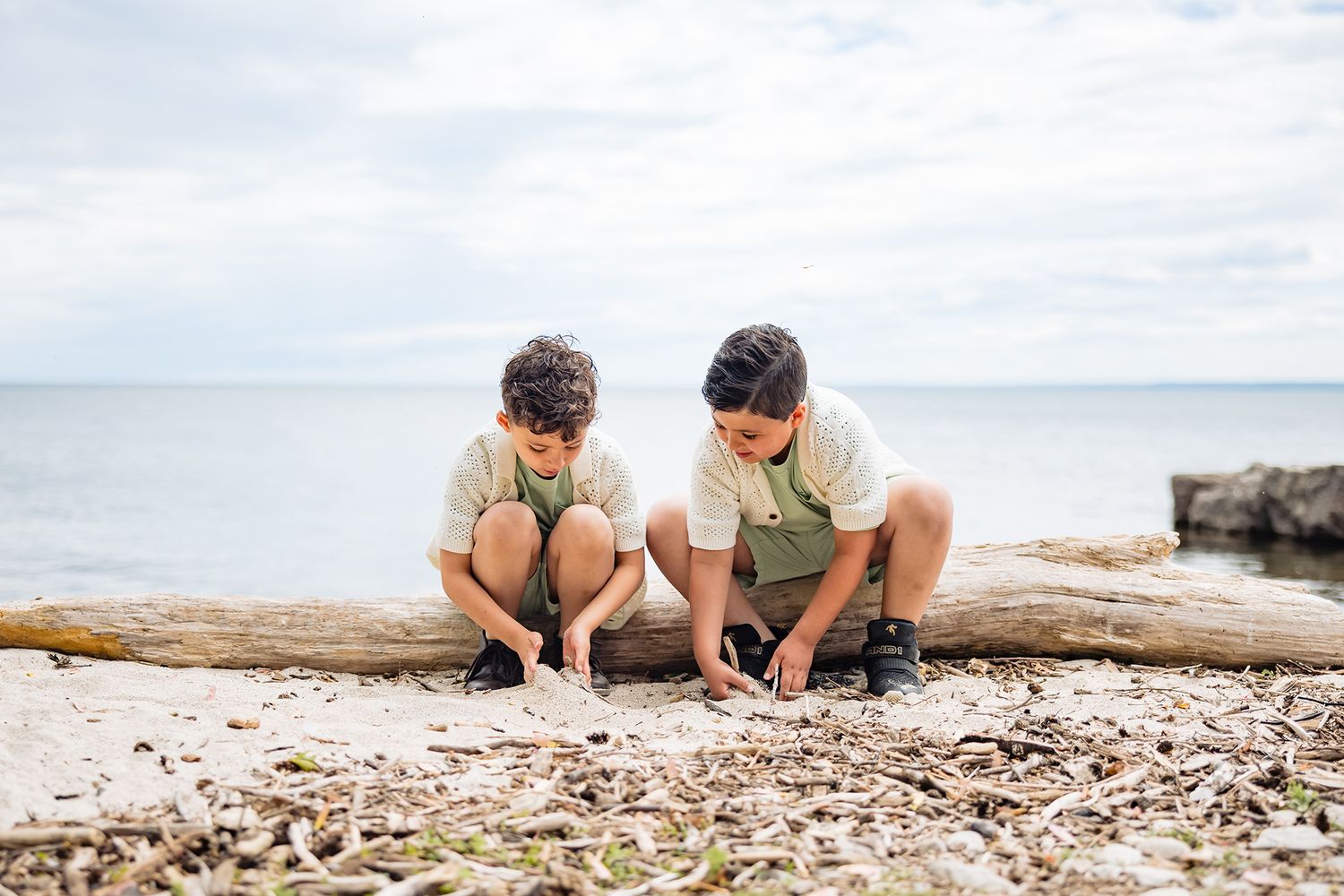 Two children crouched on a log by the ocean examining items on the beach.