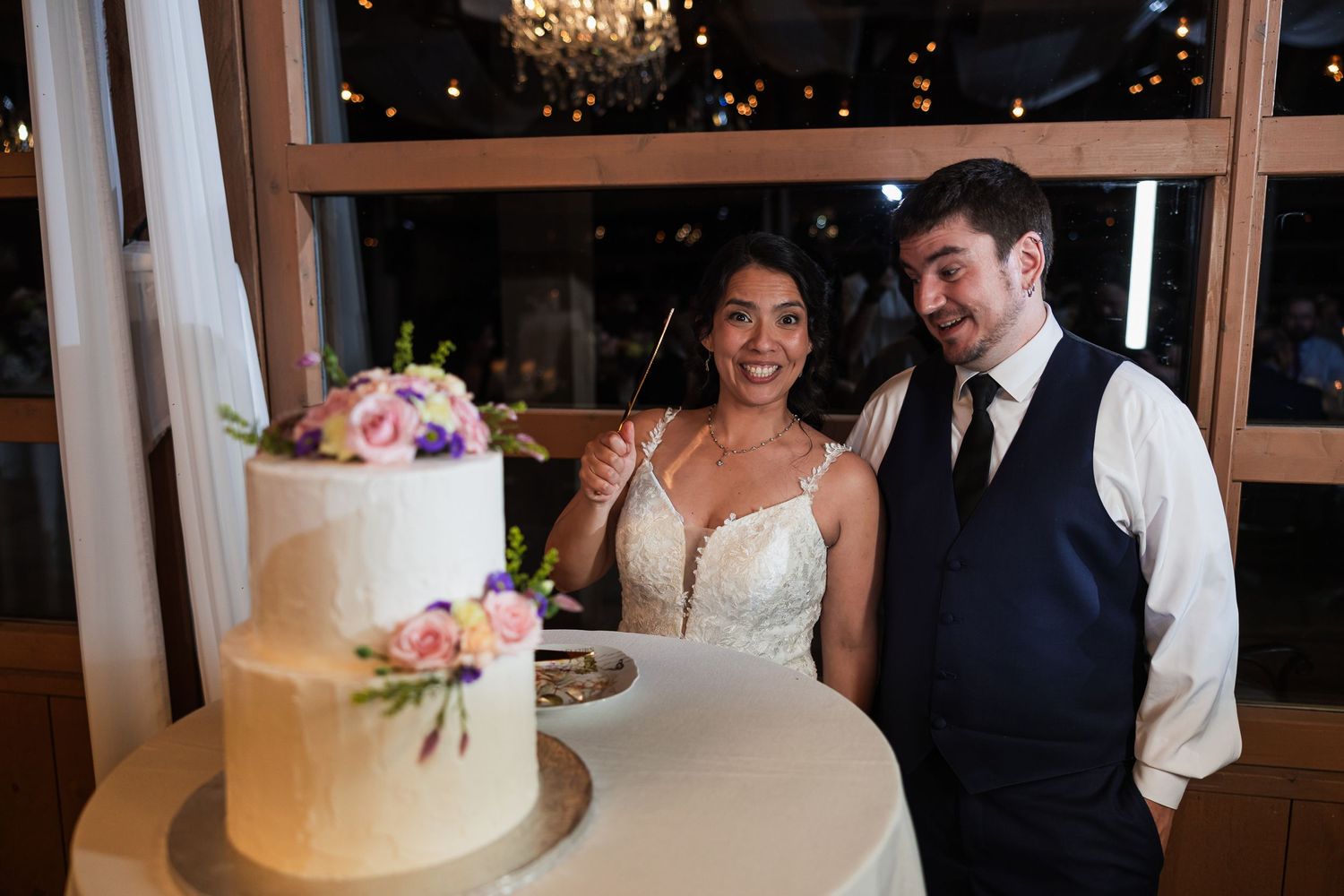 Bride and groom smiling while cutting their elegant two-tier white wedding cake decorated with colorful flowers.