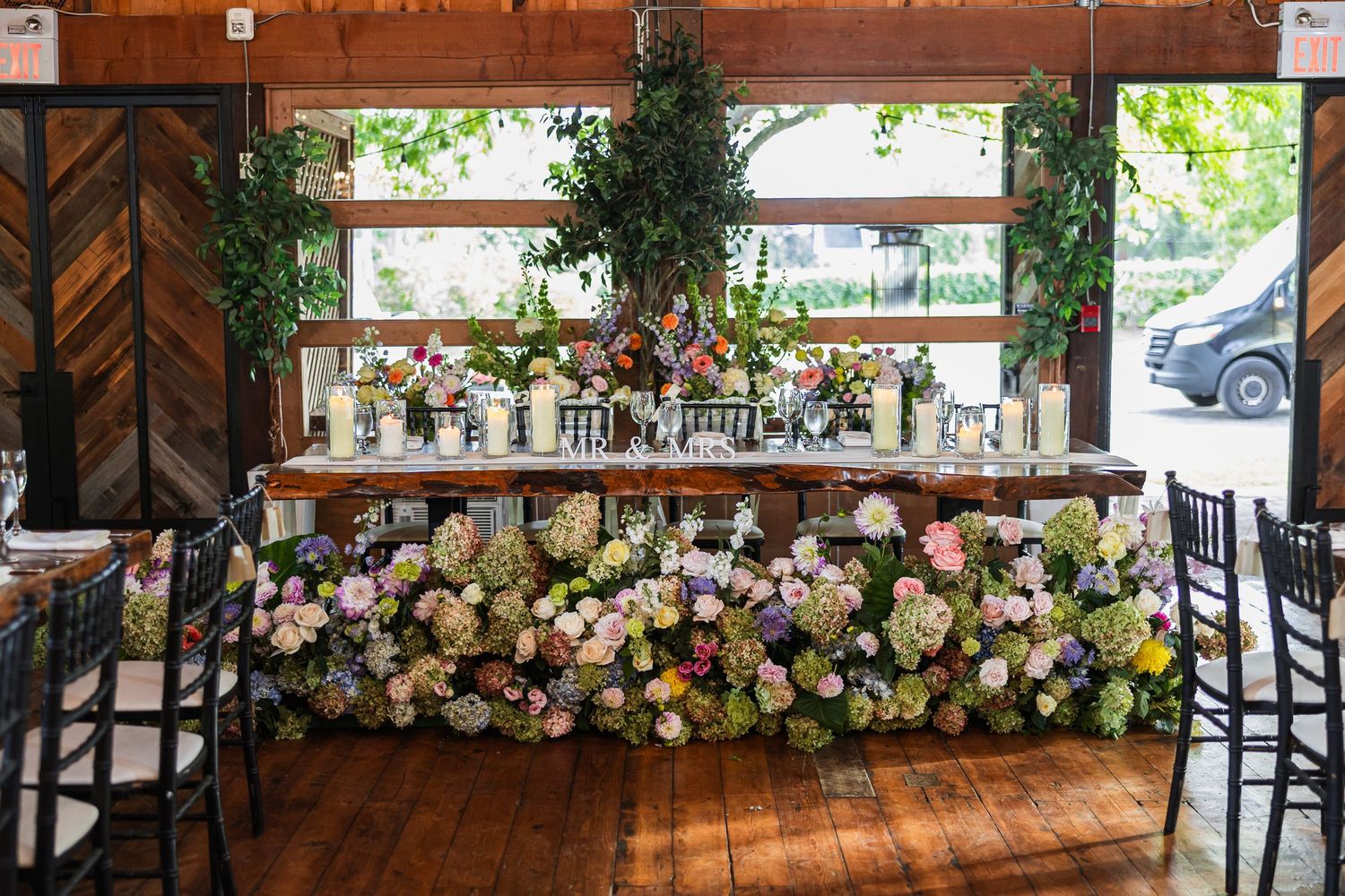 Elegant wedding head table adorned with lush floral arrangements of pink, white, and green blooms in a rustic barn venue.