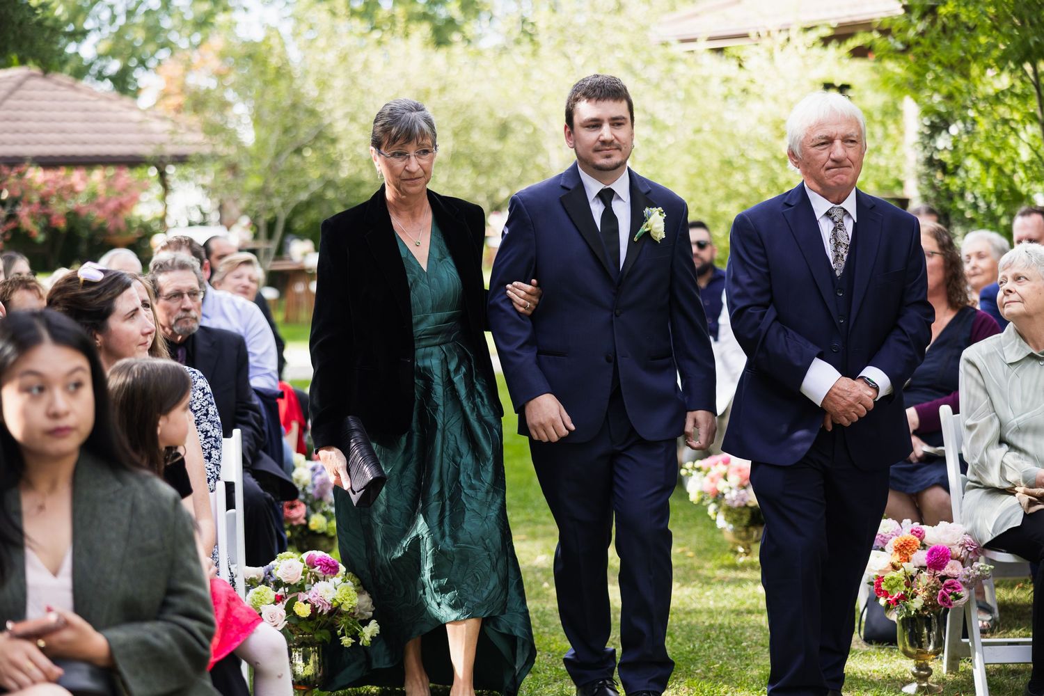 Groom walking down the aisle escorted by parents at an elegant outdoor garden wedding ceremony.