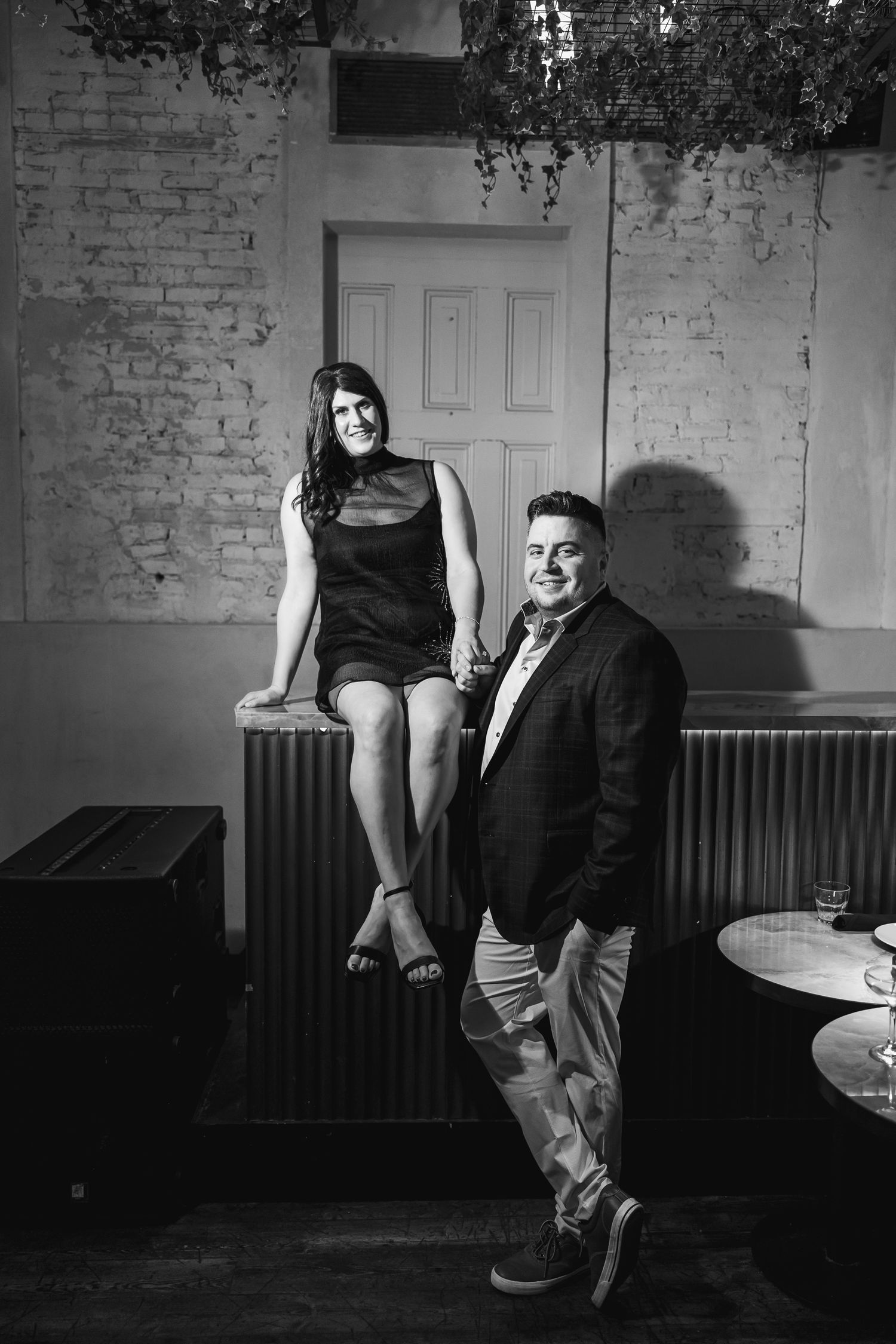 Black and white photo of a smiling couple posing in a rustic brick-walled venue, woman sitting on ledge, man standing beside her.