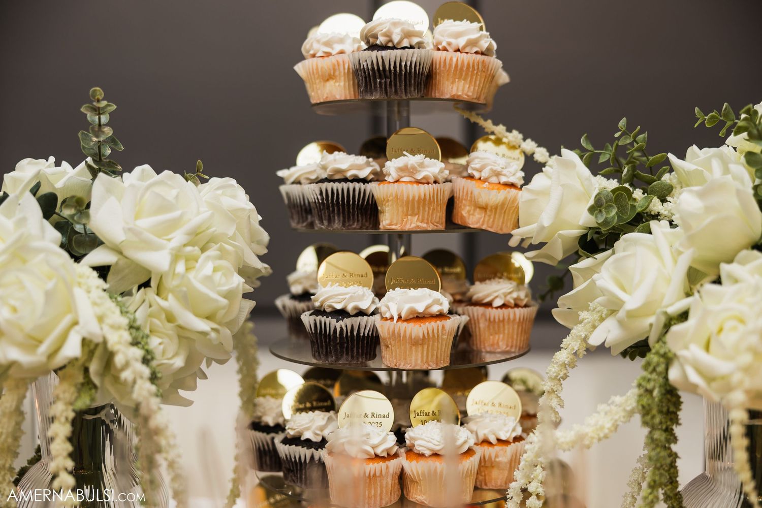 Elegant dessert display featuring cupcakes and white roses on tiered stands with golden accents.