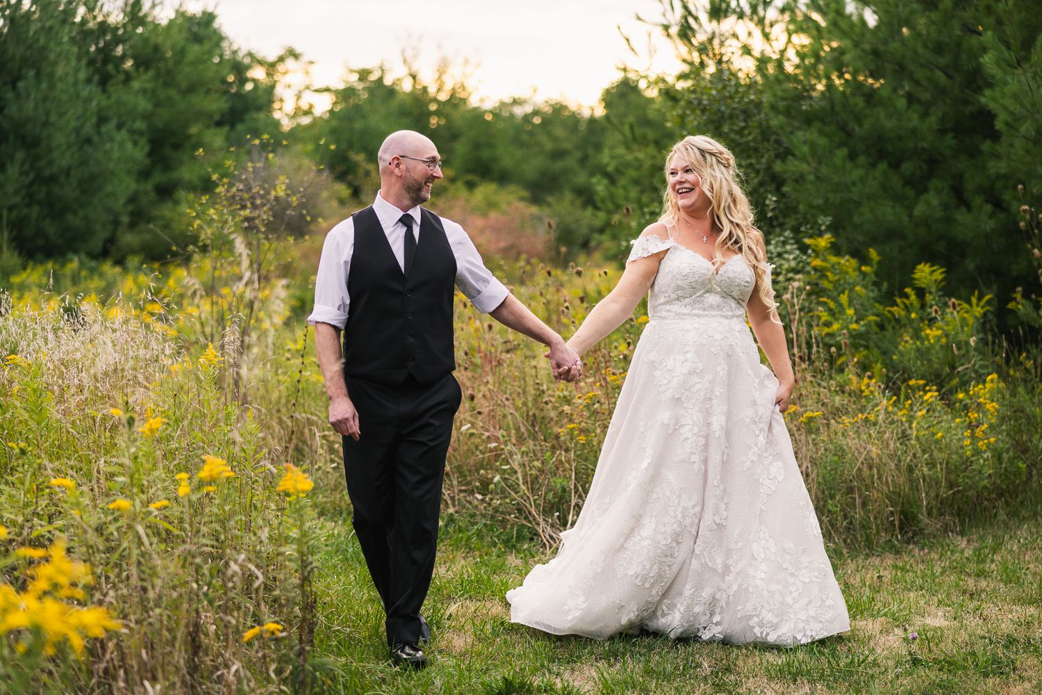 Couple walking through a sunlit meadow with yellow wildflowers at sunset during their wedding photos.