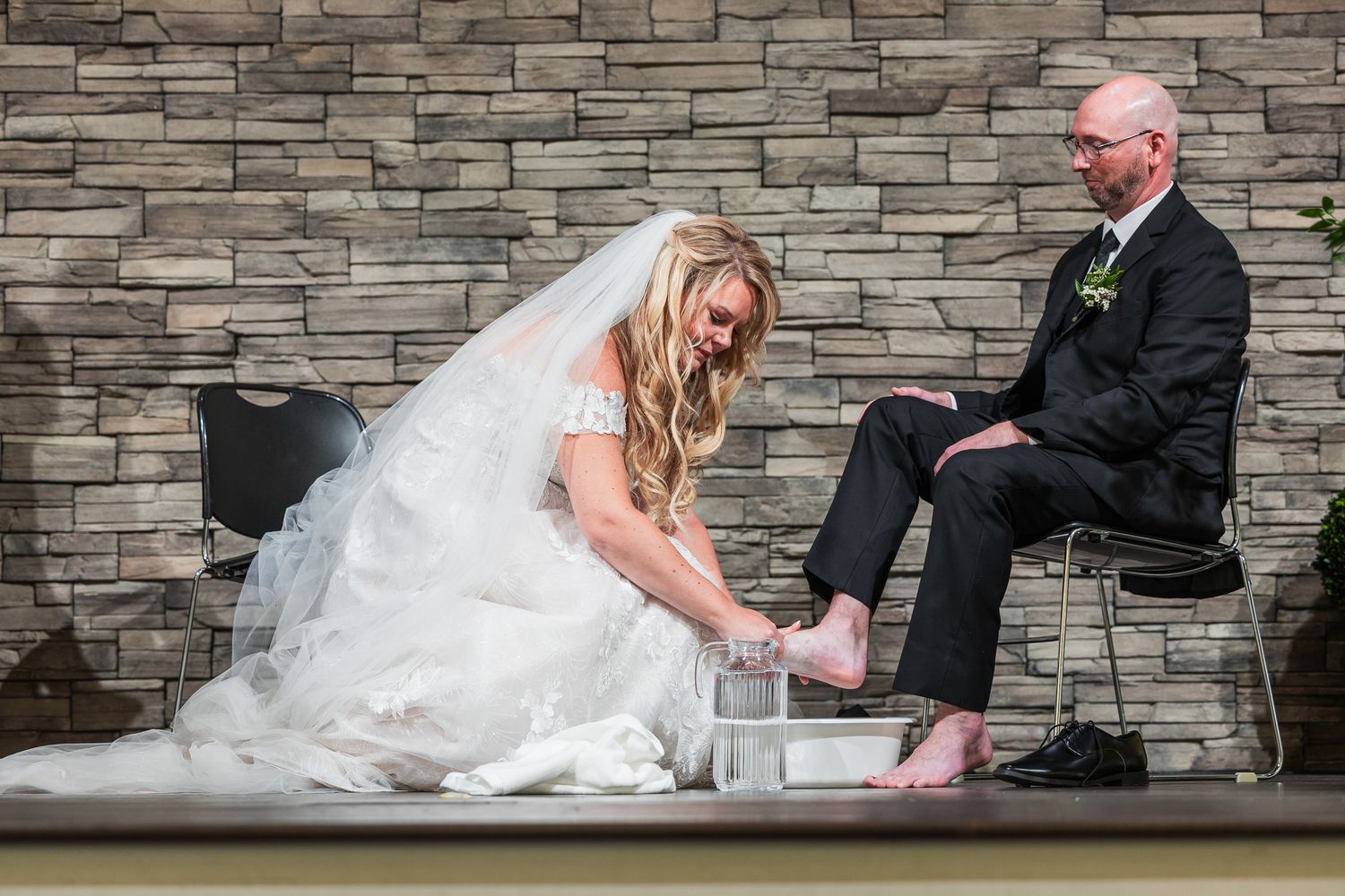 Couple sitting on stone steps share playful moments in formal wedding attire against textured wall backdrop.